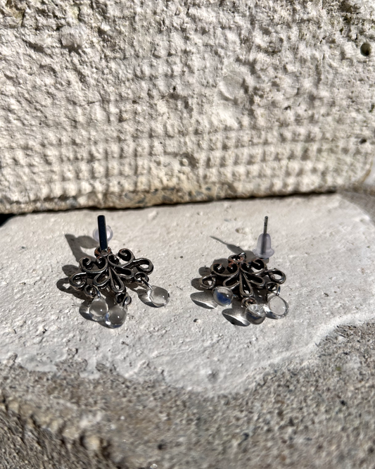 Boucles d'oreilles façonnée à partir de deux arabesques métalliques d'un vieux bracelet ottoman, montées sur des puces rectangulaires en inox hypoallergénique.
J'adore ces volutes figées dans le temps, comme une danse capturée dans le métal. Pour ajouter une touche éthérée et sophistiquée, j'ai suspendu de petites "gouttes d’eau" en verre translucide, apportant légèreté et mouvement aux boucles. Vue de profil.