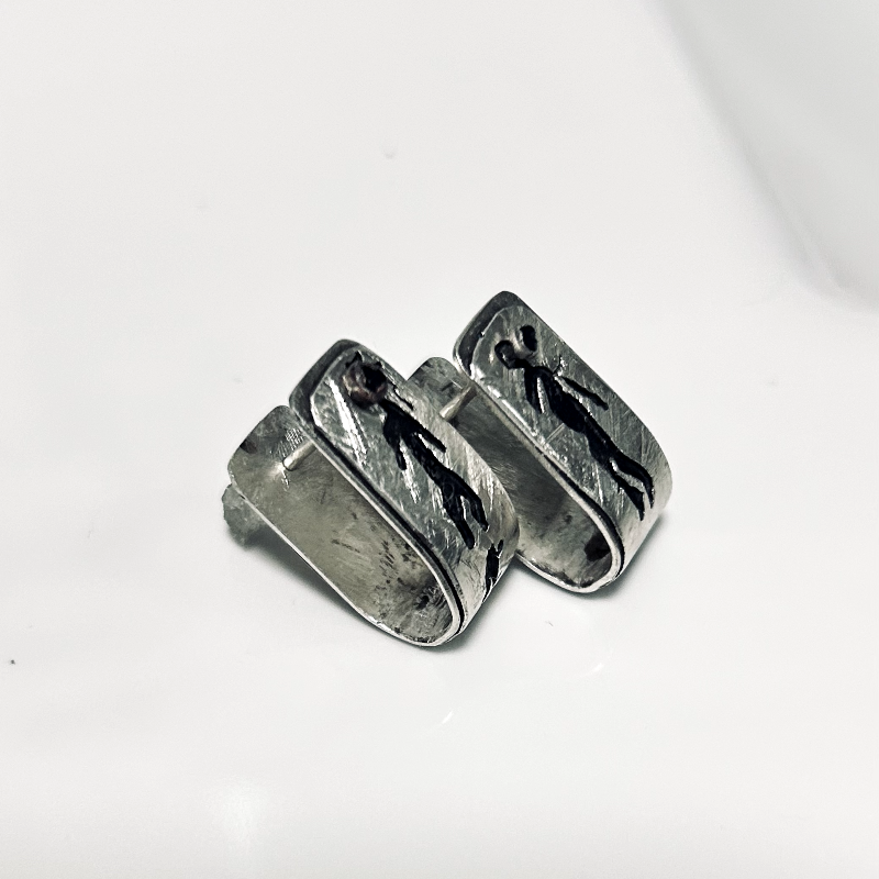 Vue de côtés sur l'attache- Boucles d'oreilles formes agrafes en argent texturés, posées sur une faïence blanche, parsemés de symboles rupestres ajourés et oxydés. Attache traversante avec poussoir silicone