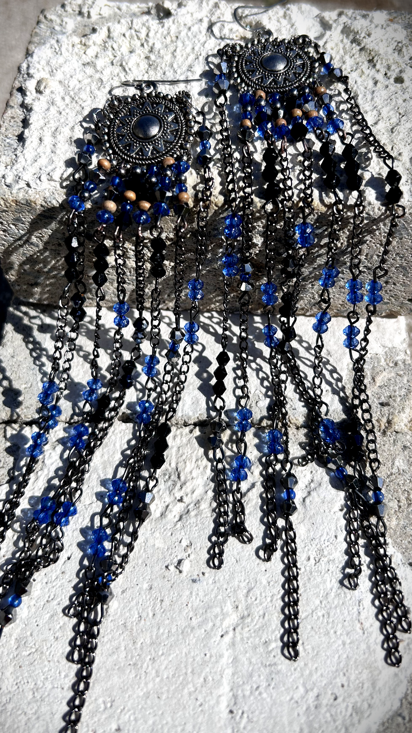 Boucles réalisées comme une évocation des tribus amérindiennes des États-Unis. Pour cela, j’ai récupéré deux maillons de bracelet, que j’ai sciés, limés, émerisés et percés afin d’y suspendre, sur des tiges inox, de longues franges mêlant chaînettes et perles. Ces franges dans les tons bleutés et noirs en symétrie inversée, apportent un équilibre parfait, et le choix de laisser le premier clou de tige libre permet un mouvement libre à chaque mouvement de tête.