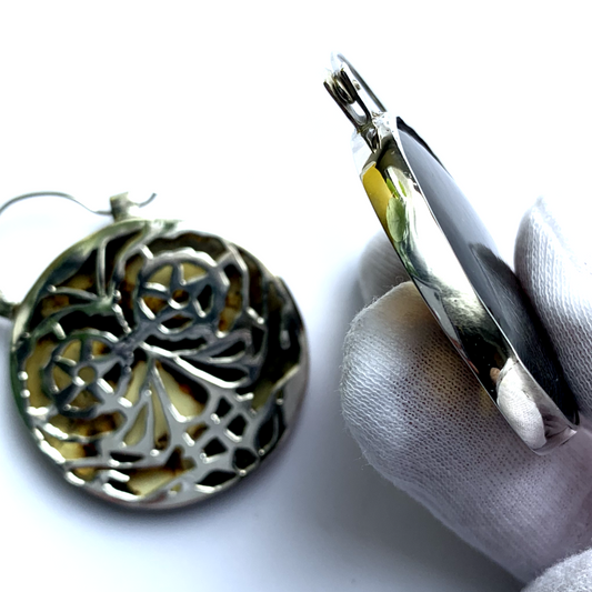 Boucles en argent de 4,5 cm de diamètre chacune, sur 6 cm de hauteur, serties de deux ambres provenant des deux veines différentes d'une mine au Chiapas, ce qui donne une particularité à cette paire de boucles harmonieuses aux couleurs variées. Le dos des sertis en argent est délicatement ajouré sur des motifs de masque illustrant le jour de la fête mexicaine Día de los Muertos, cette célébration vibrante de la vie et de la mémoire. Vues de dos côté Masque Calaveras et de profil.