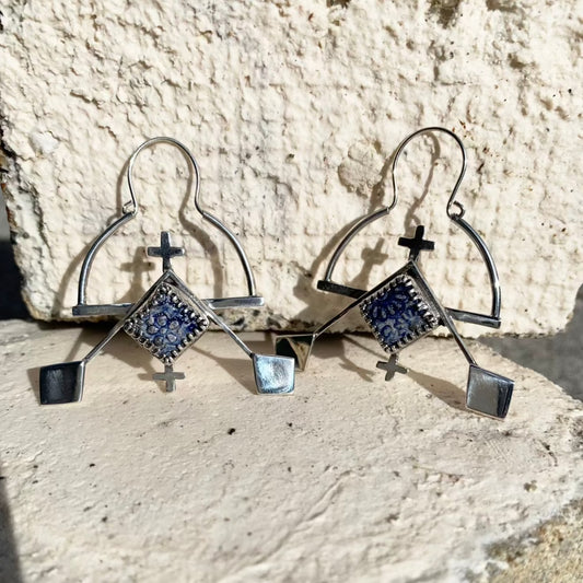 Boucles d'oreilles en argent composées d'une demi-créole en bas de laquelle est soudée une barre. Ici se trouve soudé un serti clos en argent avec une céramique raku bleue irisée carrée, encadré de croix berbères en argent. Le long des cotés du sertis sont soudés deux tiges fines en argent, où s'accrochent les deux losanges de la féminité berbères, cet ensemble symbolisant des rames au soleil couchant. Attaches par ouvertures au centre des créoles, crochets s'insérant dans le tube de cette créole.