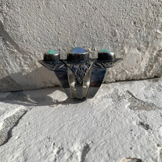 Bague composée de 3 joncs soudés et gravés de symboles berbères (serpent lunaire, grains et gâteau de miel), sertie de trois opales Welo éthiopiennes. Le tout représentent la lune en harmonie avec le serpent lunaire, illustrant une syzygie – un moment où trois corps célestes sont alignés, reliant les cycles de la lune et de la mue du serpent. Opales laiteuses aux reflets rose, jaune et bleu selon l'orientation du soleil. Vue de profil.