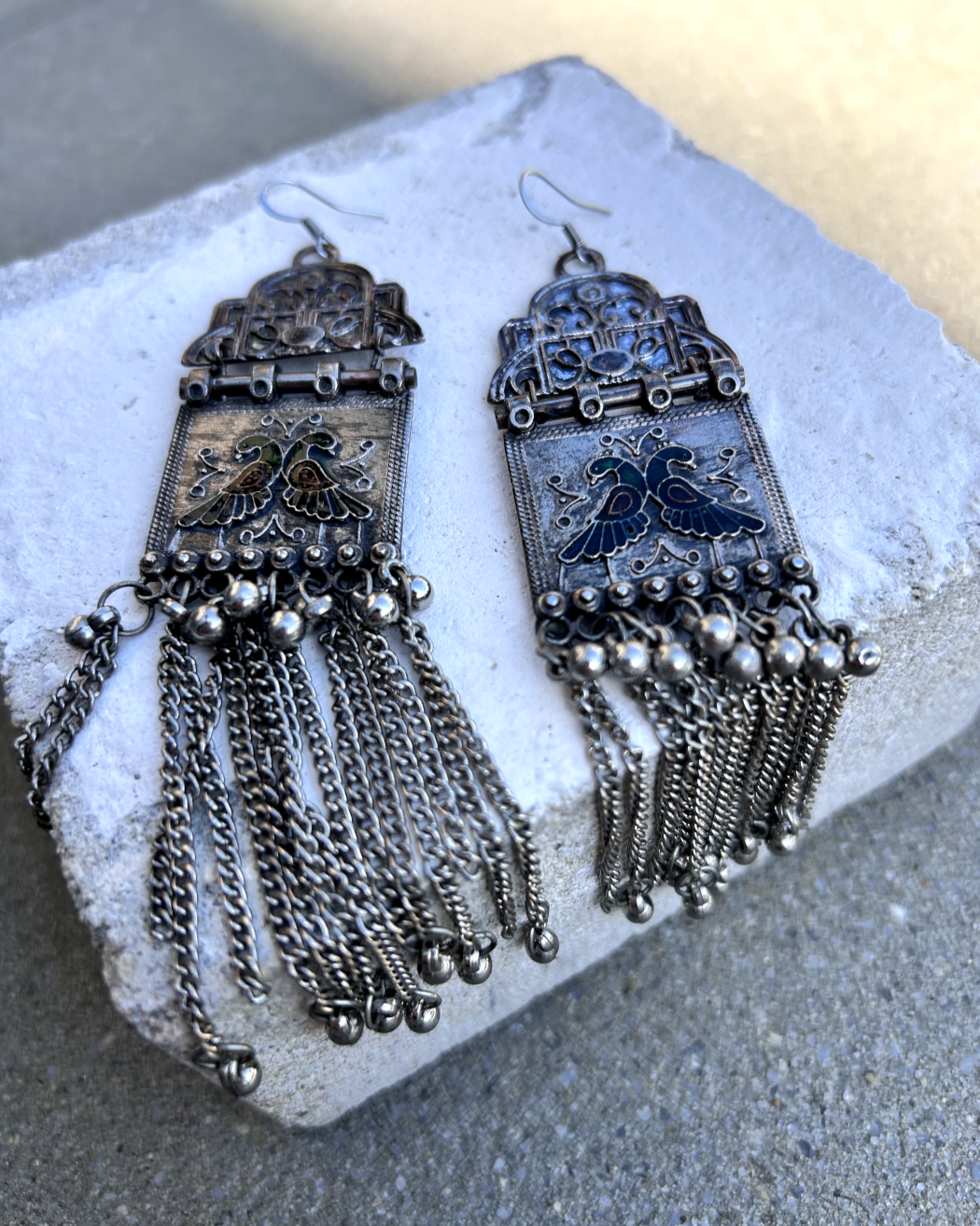 Boucles sur crochets inox, composées d'un bout de collier en alcove, auquel est supendu un tube acier d'un vieux collier qui sert de point de roulis à un carré de métal supendu, lui-même décoré de deux paons en cloisonné et pâte de verre. J'ai uspendu des pampilles récupéré sur un vieux collier indien.  je les ai noirci à la peinture métal, brossé à la brosse acier. Elles sont protégé à la cire. Je voulais  créer des boucles d'un autre âge, d'inspiration indienne avec deux paons s'embrassant sous l'alcove. 