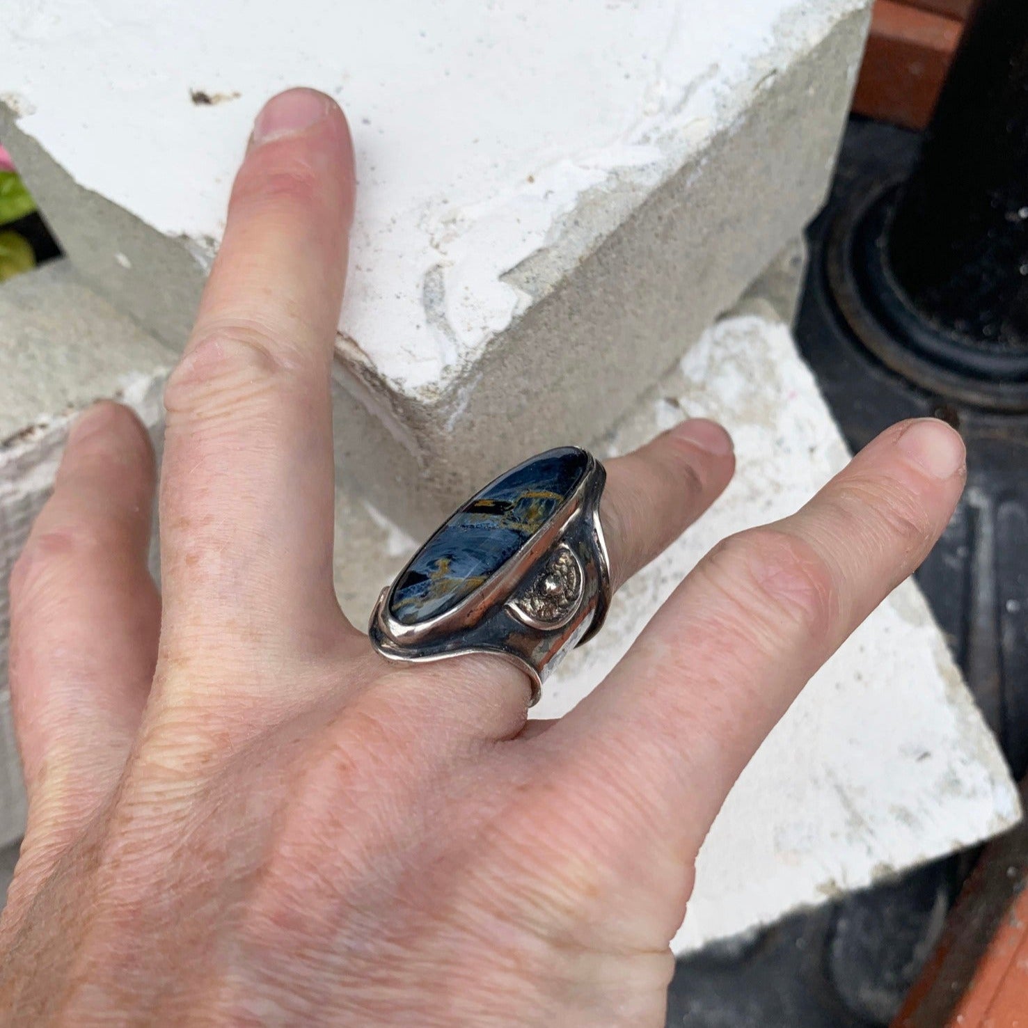 Bague réglable en argent avec un large aplat sur le dessus serti d'une pietersite bleu nuit, entourée de deux symboles de demi-lune berbère texturé. Chaque demi-lune de part et d'autres de la pierre possède une bille centrale qui vient compléter ce symbole porte bonheur. Argent oxydé tons bleus. Vue de quart.