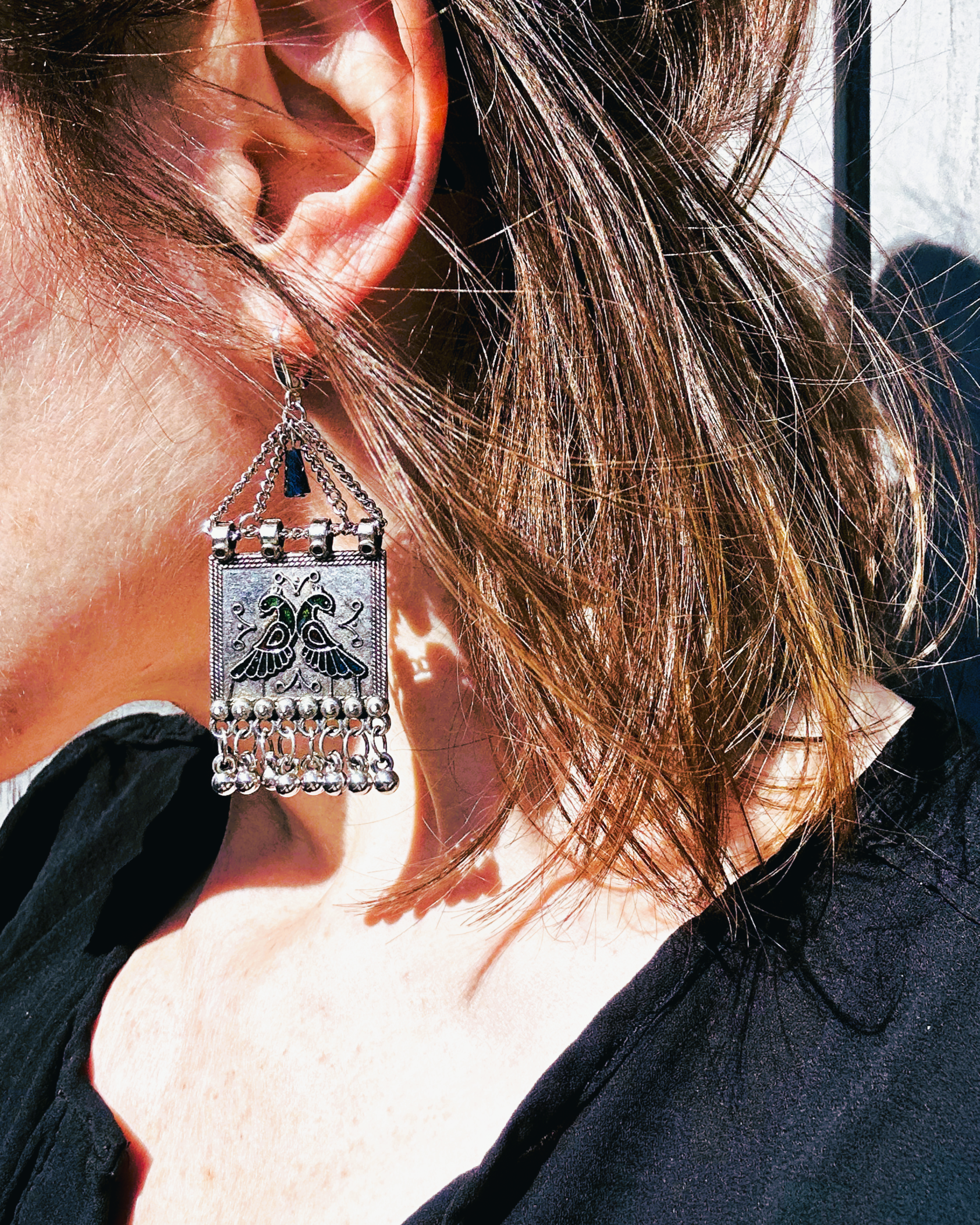 Boucles sur crochets sécurité en argent 950, composées d'un jeu de chainettes auxquelles  est suspendu une plaque de métal décoré de deux paons (cloisonné et pâte de verre). J'ai suspendu des pampilles récupéré sur un vieux collier indien.  jApreès avoir retiré la rouille, je les ai noirci à la peinture métal, puis brossé à la brosse acier. Elles sont protégé à la cire. Je voulais  créer d'inspiration indienne avec ce couple de paon aux couleurs chatoyantes vertes, bleues et rouges. Vue portée au soleil.