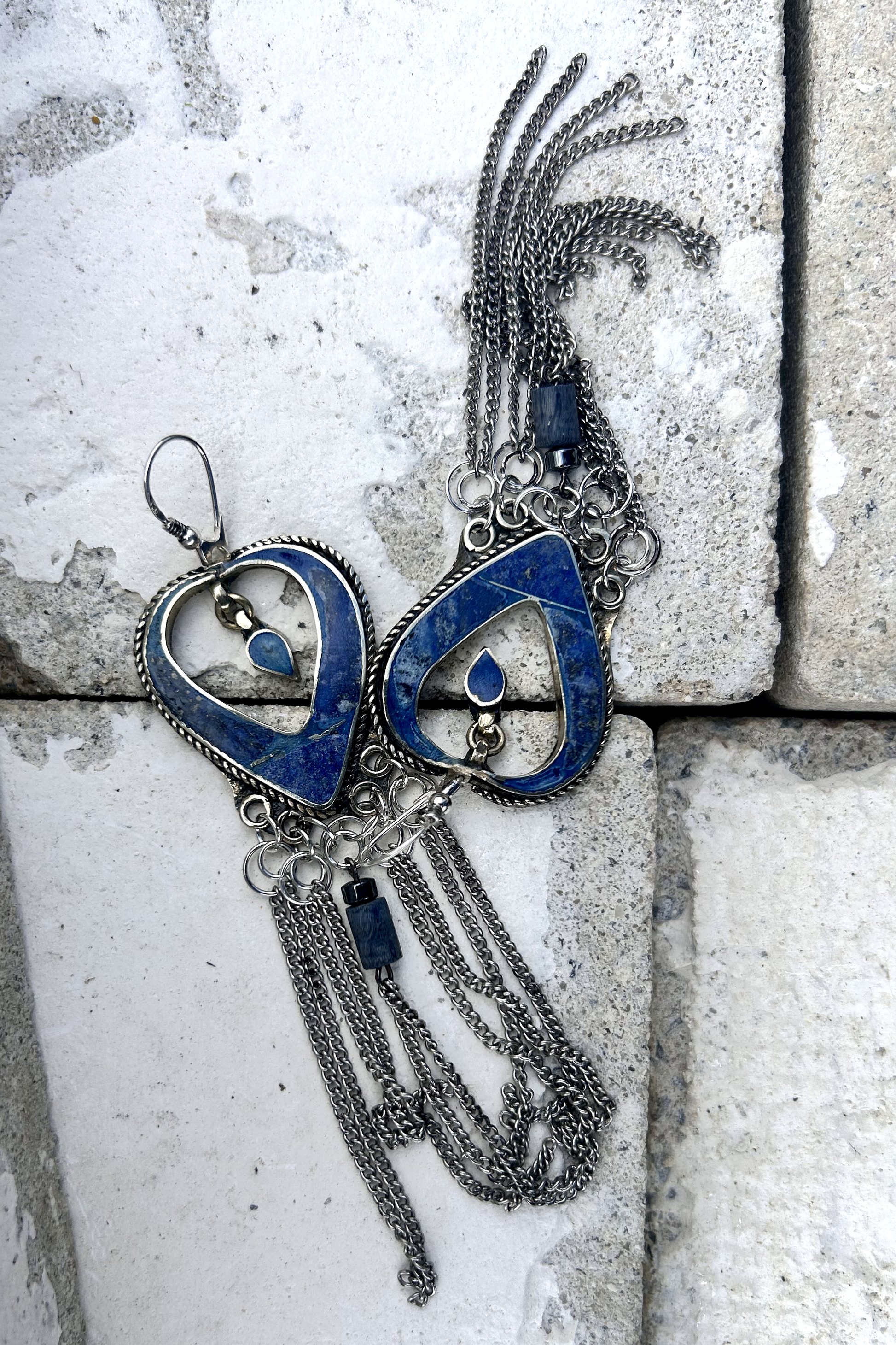 Boucles d'oreilles longues réalisées à partir de deux pendentif coeur d'un vieux collier d'Essaouira, sciées, limées, émerisé, polies et entièrement vernies, y comris la pate de verre bleu sertie dans le cloisonné. Dessous j'ai gardé qelques attaches pour construire un jeu d'anneau en inox auxquels j'ai supsendu des chaines et deux perles (une hématite, et une perle en pierre bleue). Au centre du coeur j'ai suspendu un petit charme du colllier pour illustrer les larmes du coeur. Vue dedessus, à plat.