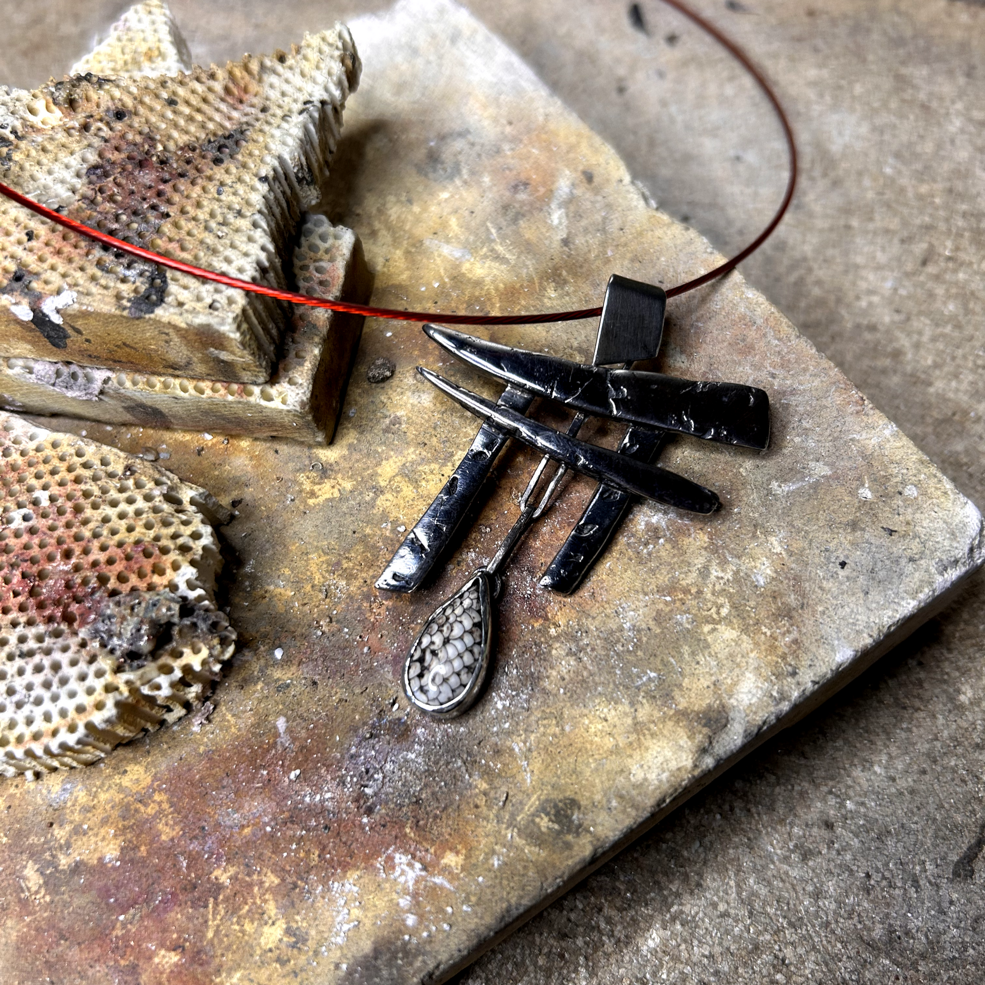 Vue de profil: pendentif réalisé à partir de débris d'argent assemblés et compressés, sculptés en Torii -portail japonais d'accès au monde des esprits. Au centre de a porte est suspendu une pierre de fossile serpent sertie.