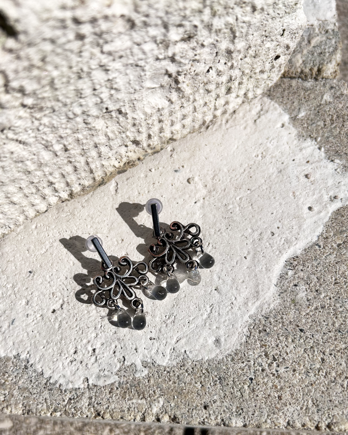 Boucles d'oreilles façonnée à partir de deux arabesques métalliques d'un vieux bracelet ottoman, montées sur des puces rectangulaires en inox hypoallergénique.

J'adore ces volutes figées dans le temps, comme une danse capturée dans le métal. Pour ajouter une touche éthérée et sophistiquée, j'ai suspendu de petites "gouttes d’eau" en verre translucide, apportant légèreté et mouvement aux boucles. Vue de dessus