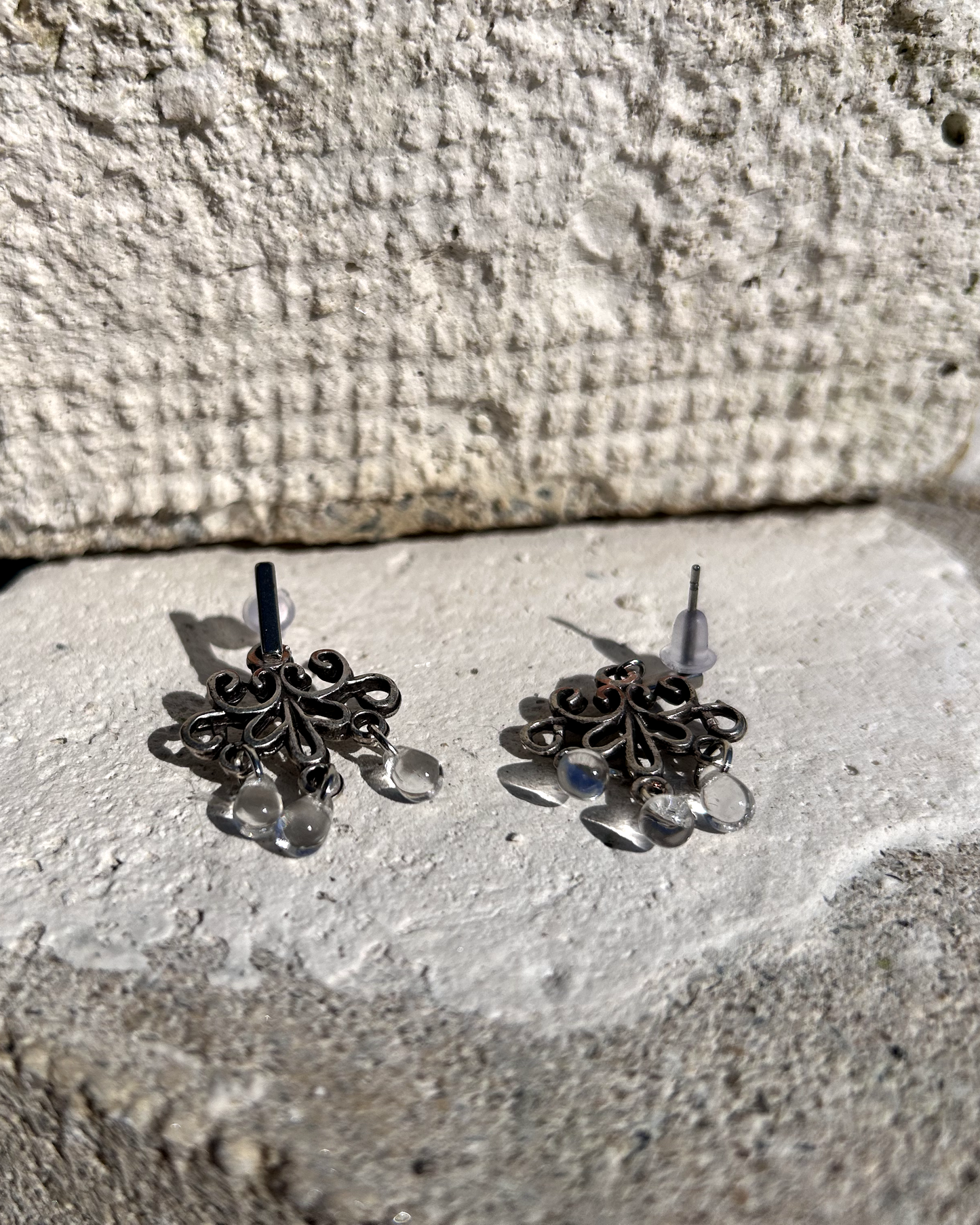 Boucles d'oreilles façonnée à partir de deux arabesques métalliques d'un vieux bracelet ottoman, montées sur des puces rectangulaires en inox hypoallergénique.

J'adore ces volutes figées dans le temps, comme une danse capturée dans le métal. Pour ajouter une touche éthérée et sophistiquée, j'ai suspendu de petites "gouttes d’eau" en verre translucide, apportant légèreté et mouvement aux boucles. Vue de profil.