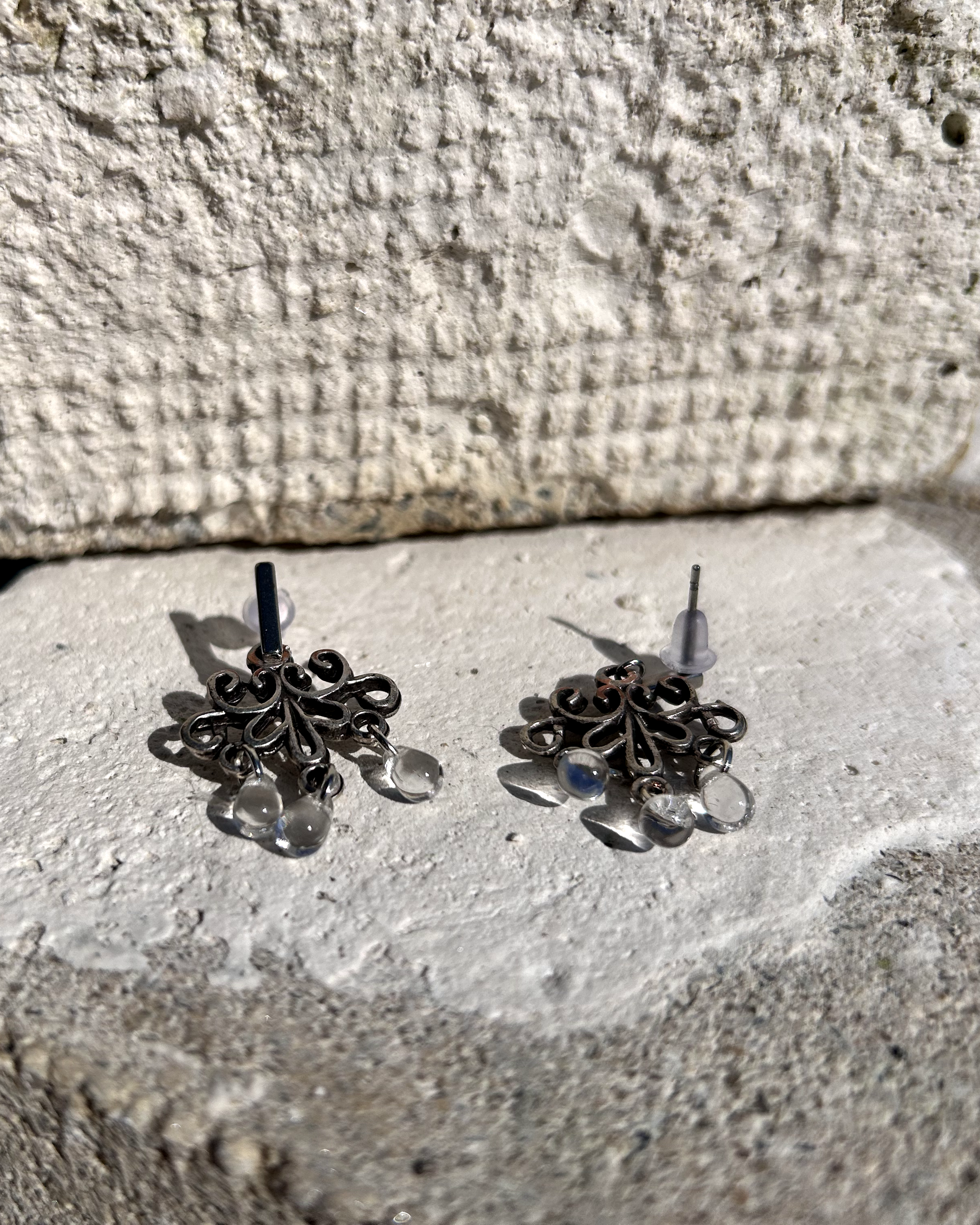 Boucles d'oreilles façonnée à partir de deux arabesques métalliques d'un vieux bracelet ottoman, montées sur des puces rectangulaires en inox hypoallergénique.

J'adore ces volutes figées dans le temps, comme une danse capturée dans le métal. Pour ajouter une touche éthérée et sophistiquée, j'ai suspendu de petites "gouttes d’eau" en verre translucide, apportant légèreté et mouvement aux boucles. Vue de profil.