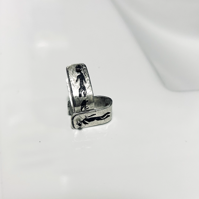 Vue de dessus- Boucles d'oreilles formes agrafes en argent texturés, posées sur une faïence blanche, parsemés de symboles rupestres ajourés et oxydés. Attache traversante avec poussoir silicone