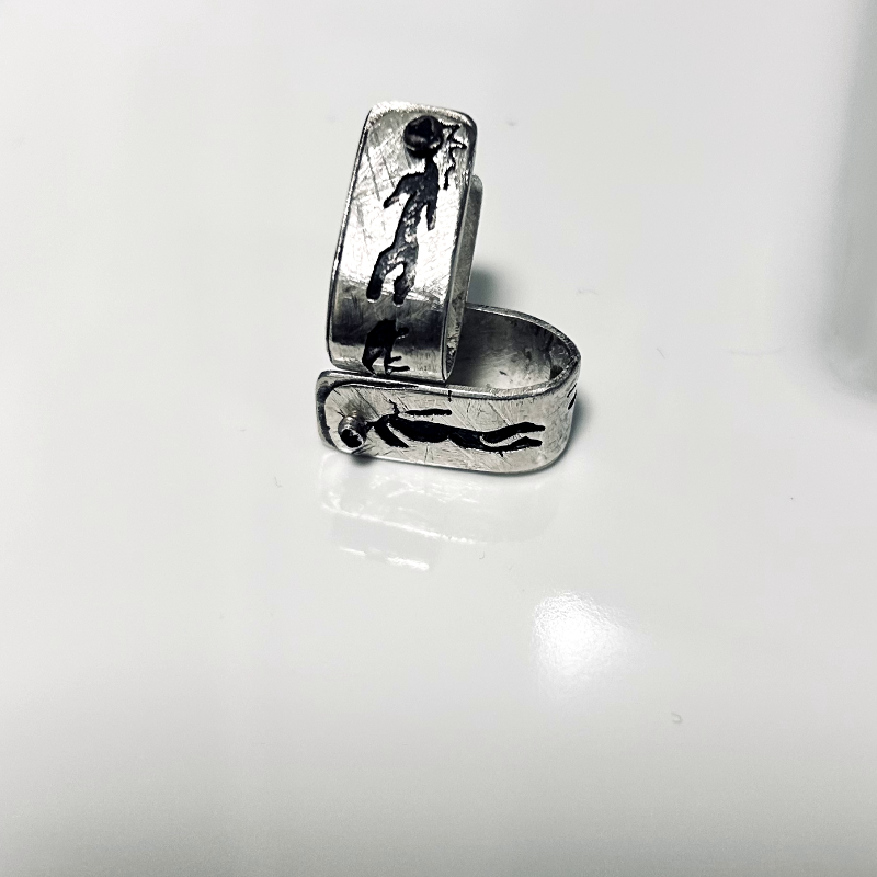 Vue de côté - Boucles d'oreilles formes agrafes en argent texturés, posées sur une faïence blanche, parsemés de symboles rupestres ajourés et oxydés (un homme et une femme) -Attache traversante avec poussoir silicone