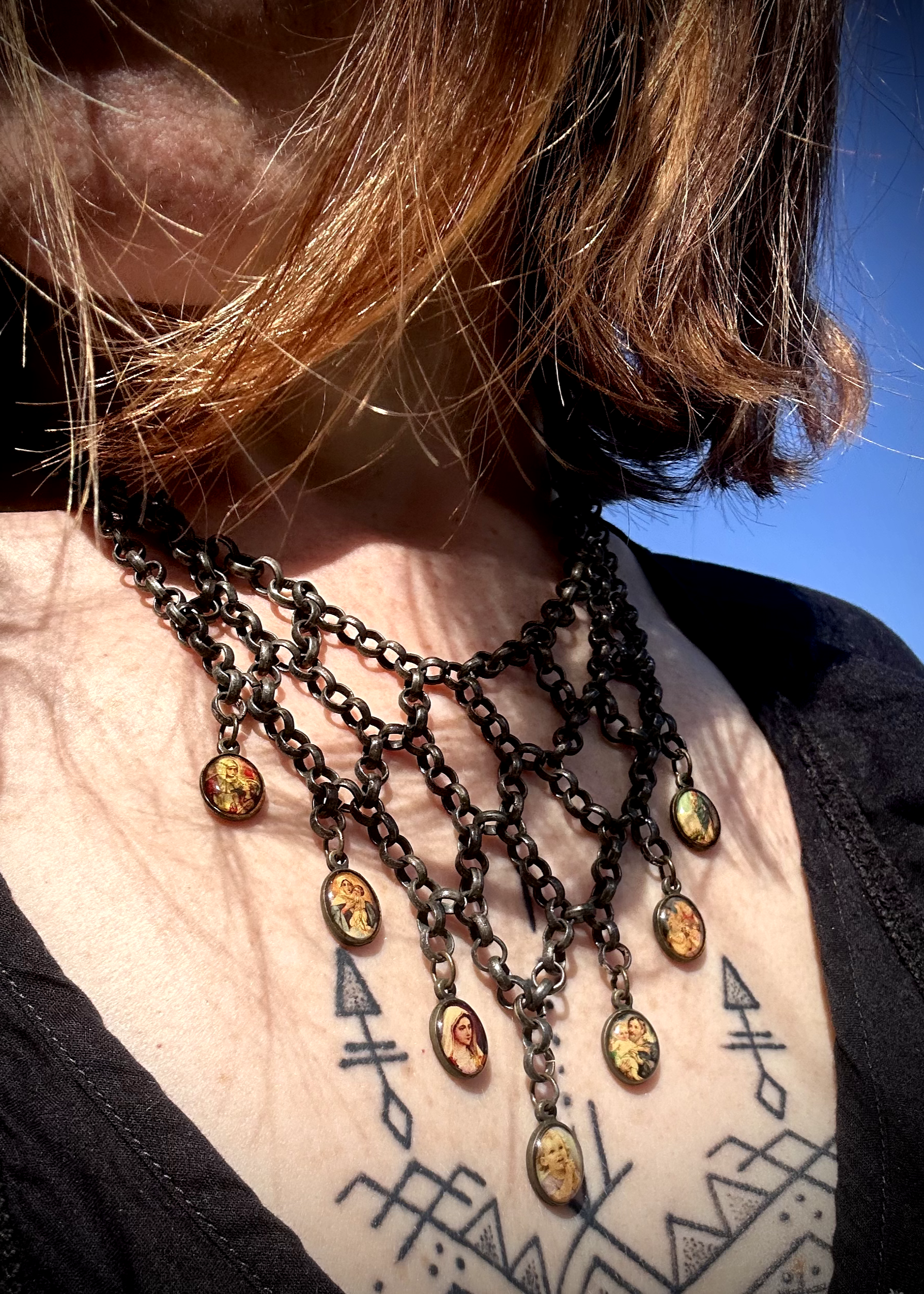 pièce unique, façonnée à partir de bijoux anciens soigneusement sélectionnés, nettoyés et réassemblés pour transmettre un message puissant:

Sept médailles religieuses vintage au fond jauni (Saints, Chérubin, Vierge), serties dans du laiton, issues d’un ancien collier de perles de culture.
Des anneaux en inox, récupérés un à un sur un collier cascade, assemblés pour créer un plastron pyramidal. Vue portée.