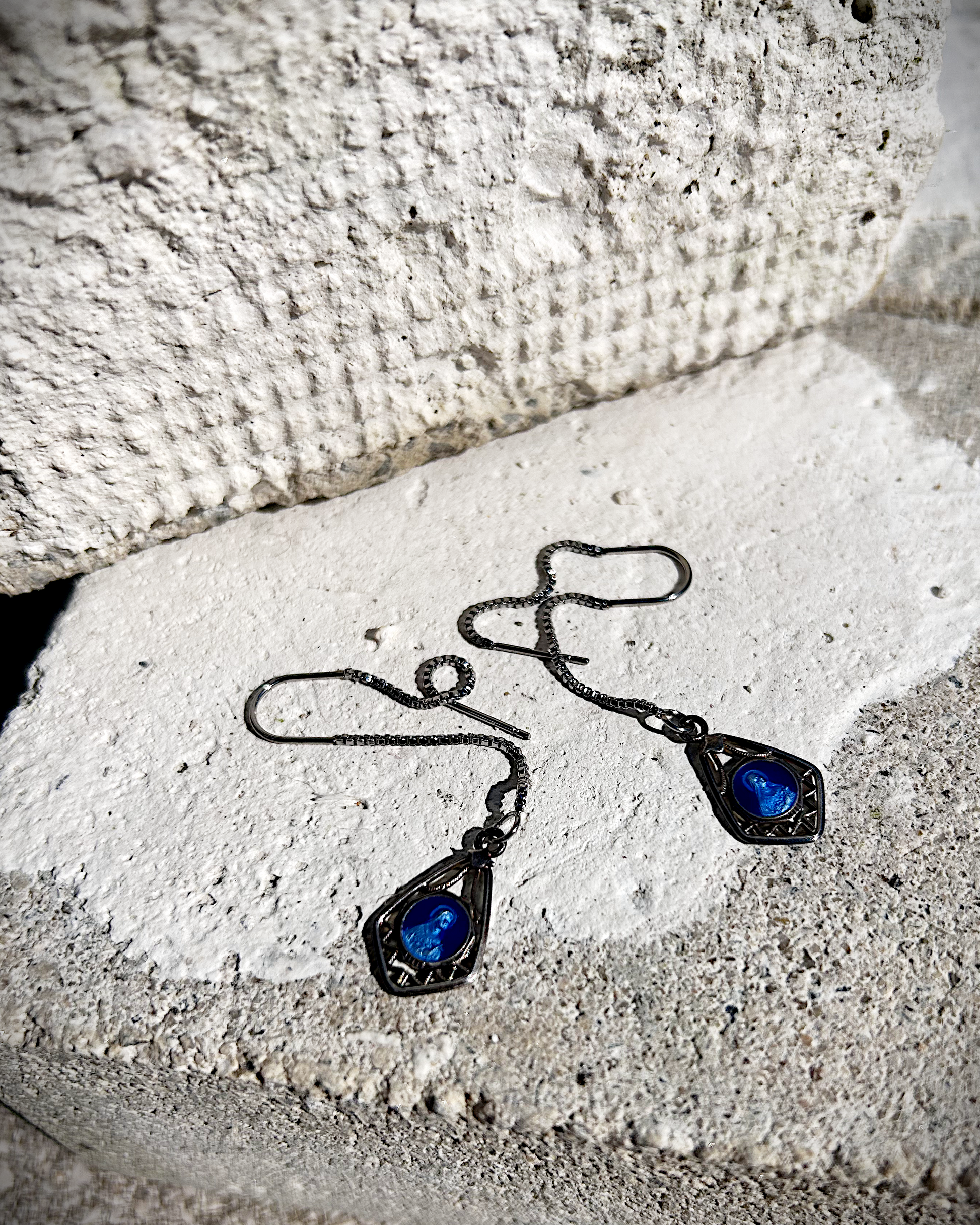 Petites boucles d’oreilles légères, composées d’une chaîne en inox hypoallergénique, suspendue à un U en inox qui traverse le lobe. À son extrémité, une médaille en métal bleu émaillée représentant la Vierge Marie est délicatement accrochée. Vue de dessus.