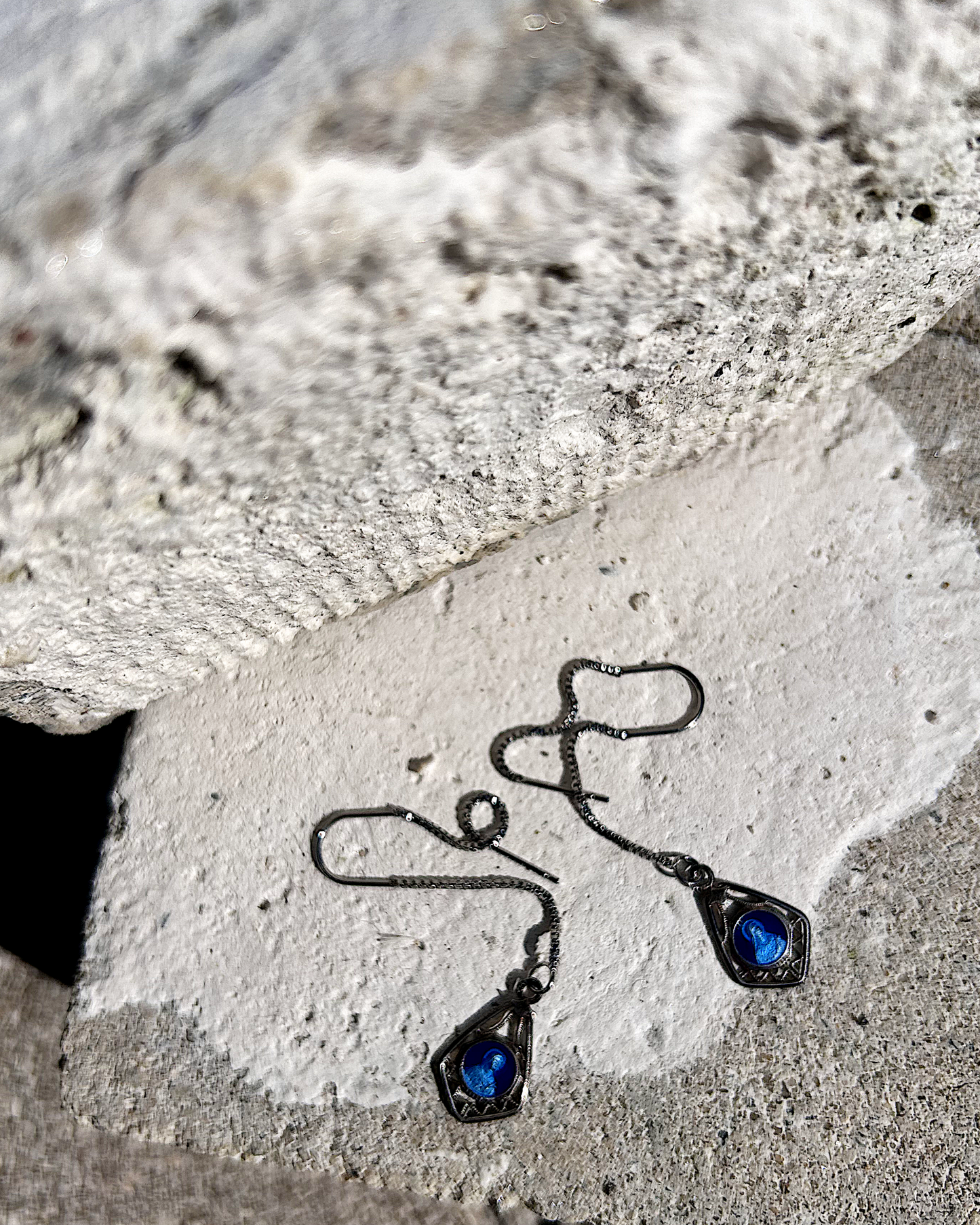 Petites boucles d’oreilles légères, composées d’une chaîne en inox hypoallergénique, suspendue à un U en inox qui traverse le lobe. À son extrémité, une médaille en métal bleu émaillée représentant la Vierge Marie est délicatement accrochée. Vue de dessus.