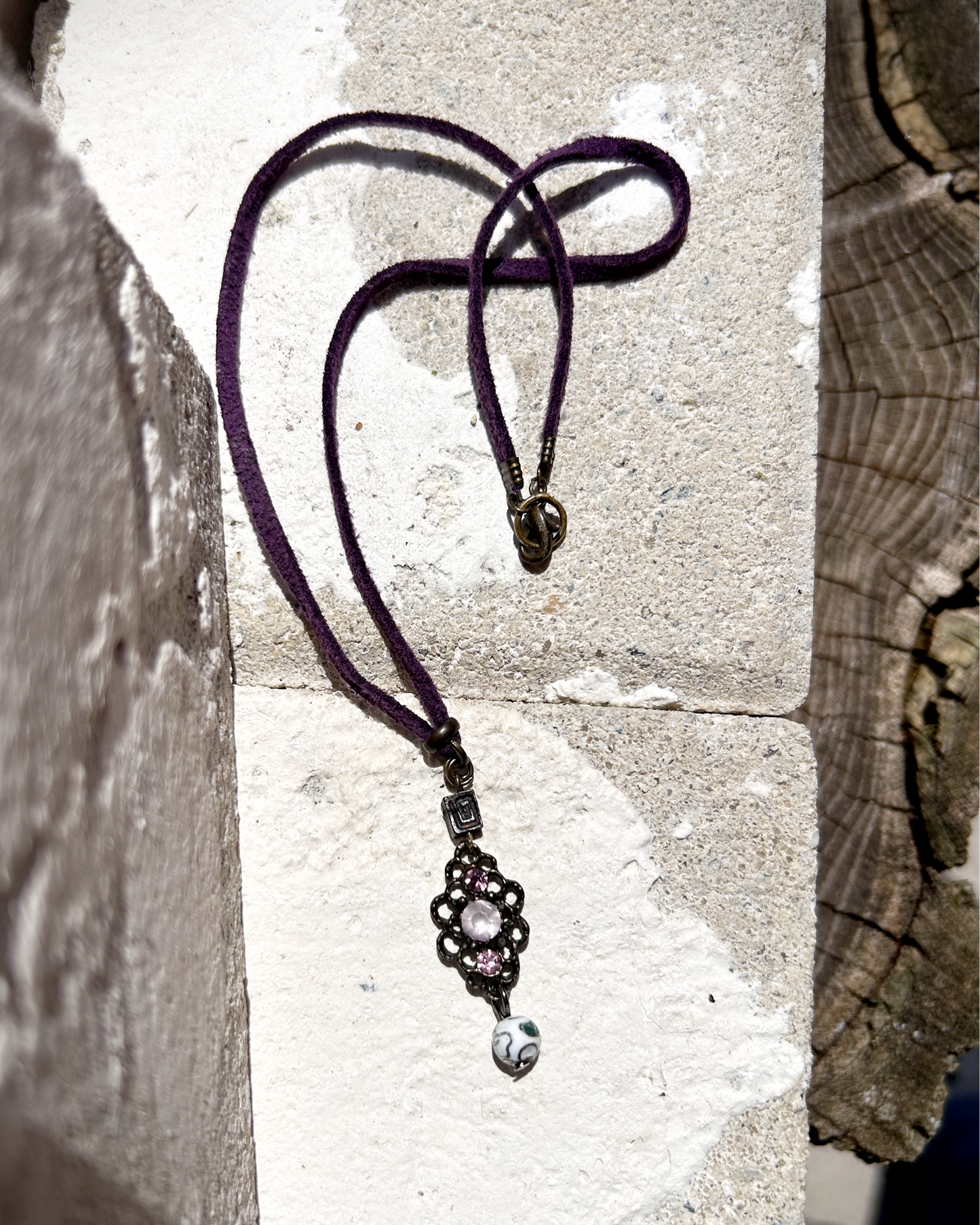 Vieux collier en suédine violette, sublimé par un charme récupéré sur un bijou ancien. J’ai ajouté deux perles : une perle japonaise en métal et une perle de verre aux tonalités délicates des cerisiers en fleurs. Tout en douceur, un brin vintage, comme un souffle léger parmi les pétales dansants des sakura. Vue posée sur une pierre.