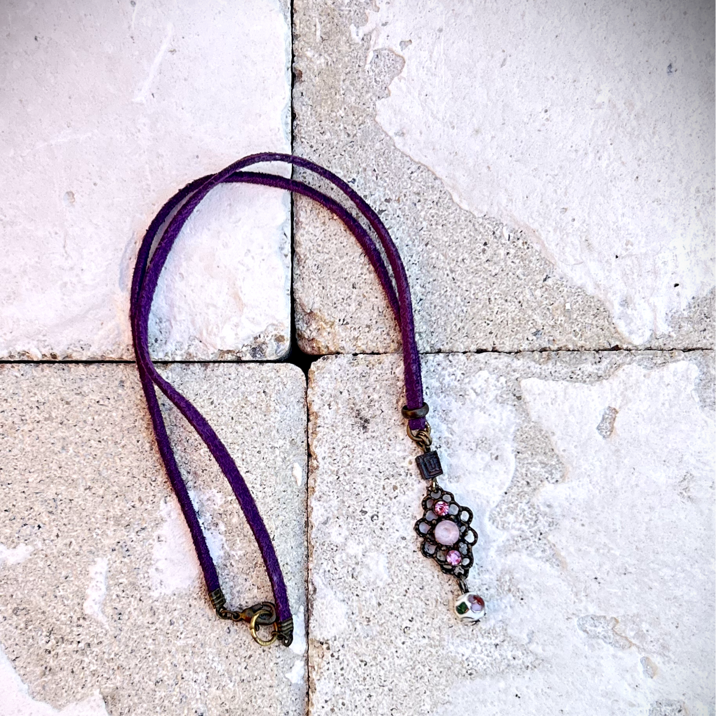 Vieux collier en suédine violette, sublimé par un charme dans les mêmes tons, récupéré sur un bijou ancien. J’ai ajouté deux perles : une perle japonaise en métal et une perle de verre aux tonalités délicates des cerisiers en fleurs. Tout en douceur, un brin vintage, comme un souffle léger parmi les pétales dansants des sakura. Vue posée sur brique au soleil.