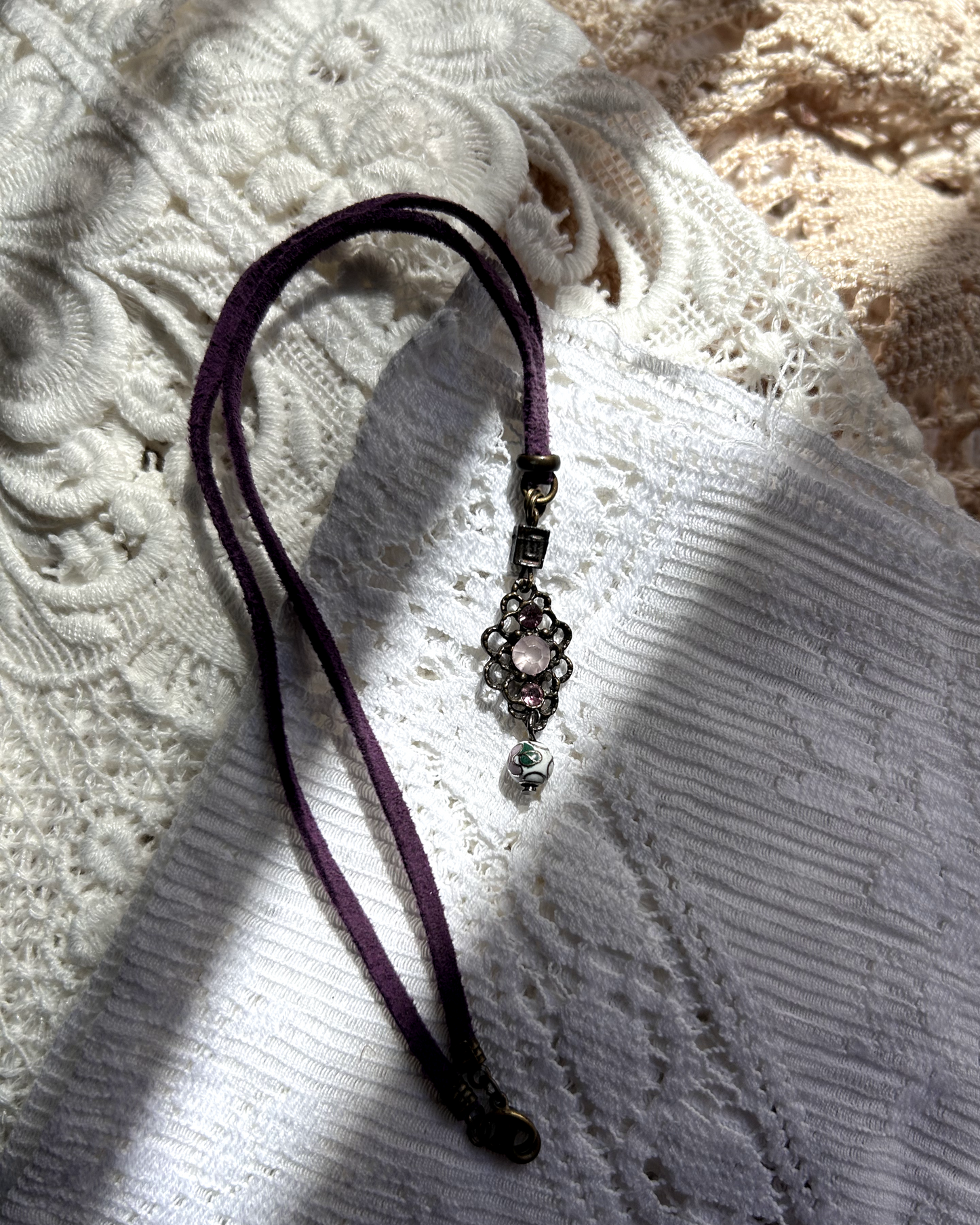 Vieux collier en suédine violette, sublimé par un charme dans les mêmes tons, récupéré sur un bijou ancien. J’ai ajouté deux perles : une perle japonaise en métal et une perle de verre aux tonalités délicates des cerisiers en fleurs. Tout en douceur, un brin vintage, comme un souffle léger parmi les pétales dansants des sakura. Vue du dessus.