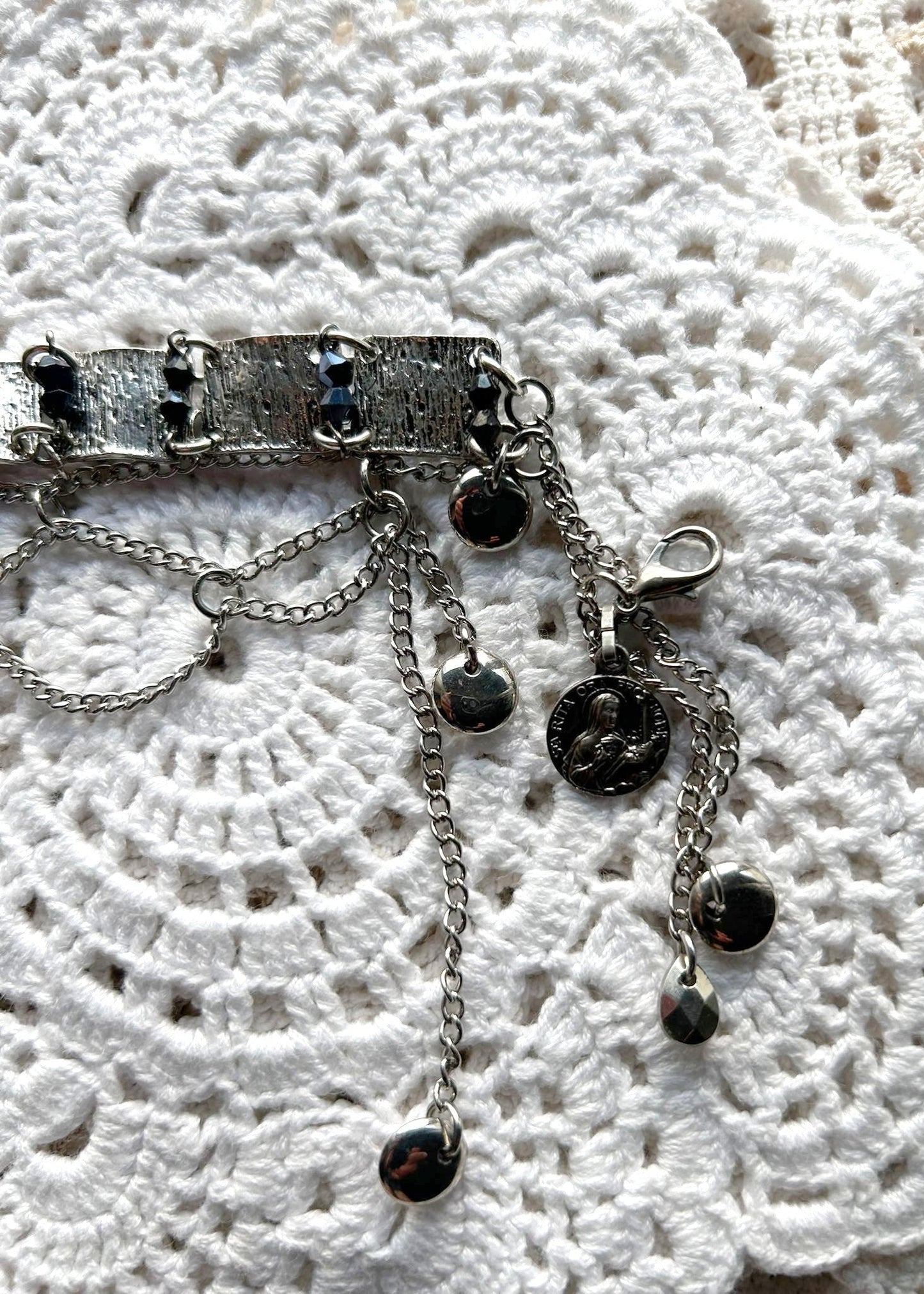 Ce bracelet est une pièce unique, façonnée avec à partir de matériaux chinés puis, soigneusement sélectionnés : Chainettes et carrés de métal récupérés sur un collier berbère, brossés puis courbés. Perles double cône losange noires et bleutés brillantes récupérés sur un collier de franges de perles, fixés sur tiges en inoxydable. Fermoir menotte. Vue de la médaille de Sainte Rita.