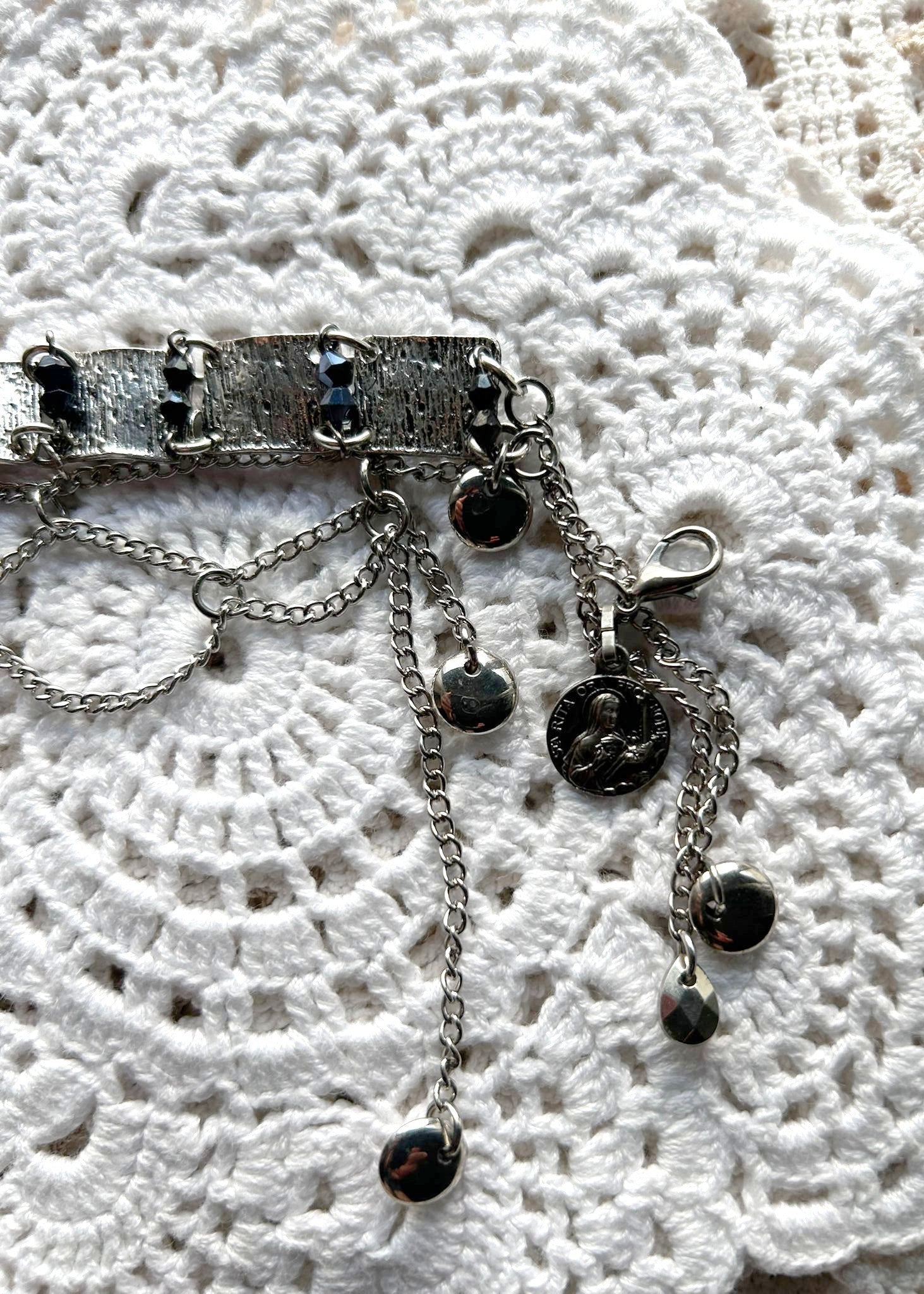 Ce bracelet est une pièce unique, façonnée avec à partir de matériaux chinés puis, soigneusement sélectionnés : Chainettes et carrés de métal récupérés sur un collier berbère, brossés puis courbés. Perles double cône losange noires et bleutés brillantes récupérés sur un collier de franges de perles, fixés sur tiges en inoxydable. Fermoir menotte. Vue de la médaille de Sainte Rita.