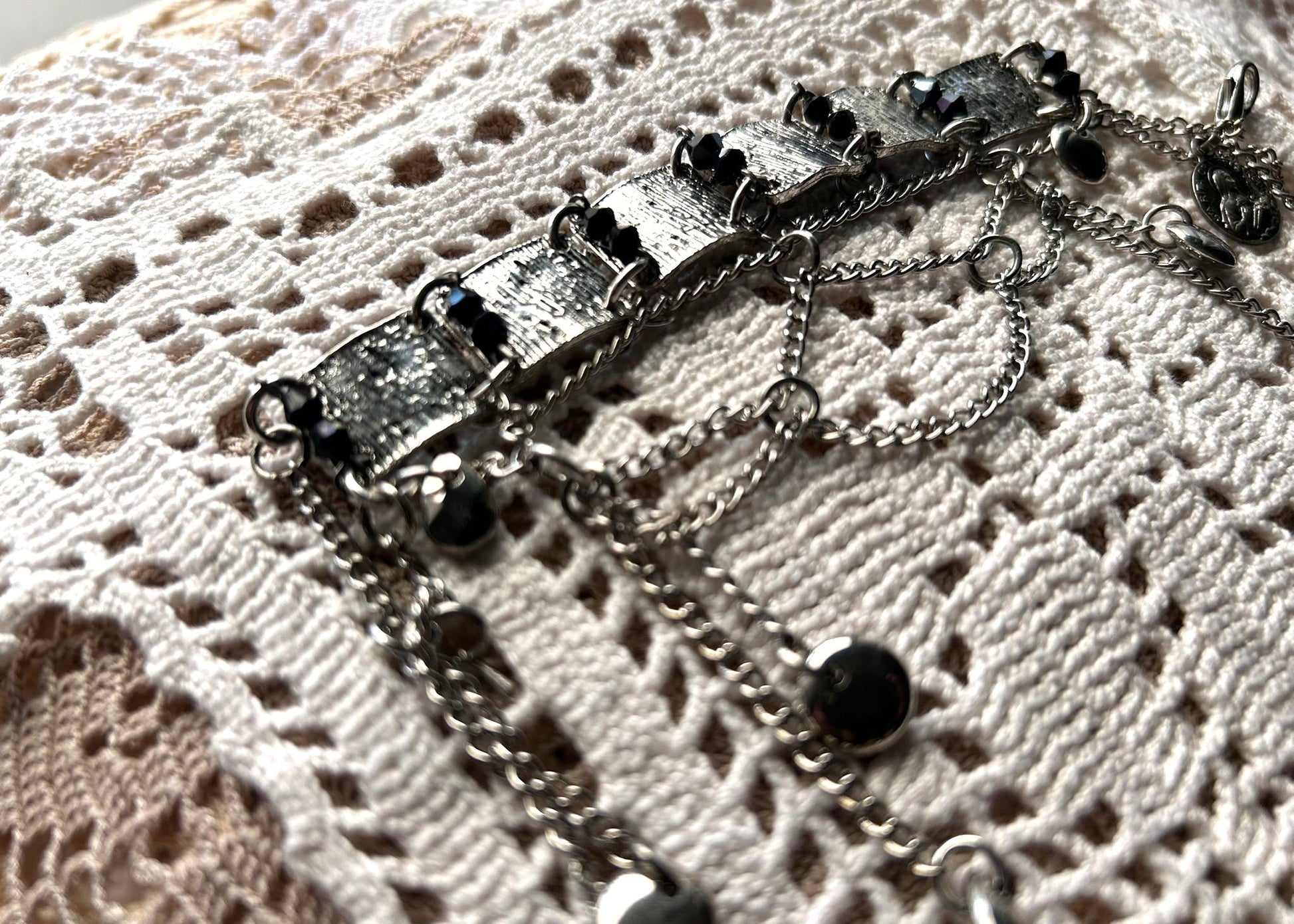 Ce bracelet est une pièce unique, façonnée avec à partir de matériaux chinés : Chainettes et carrés de métal inconnu récupérés sur un collier berbère, brossés puis courbés. Petits maillons en inox  pour alléger l’ensemble. Perles double cône losange noires et bleutés brillantes récupérés, fixés sur tiges en inoxydable. Fermoir menotte. Vue de profil posé sur dentelle au crochet.