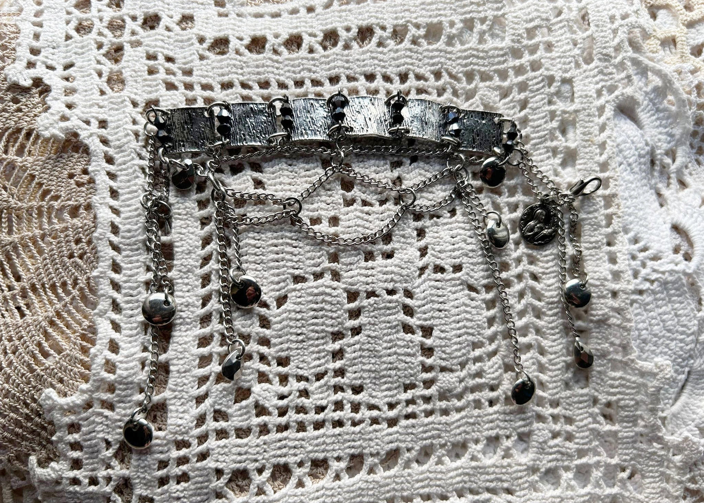 Ce bracelet est une pièce unique, façonnée avec à partir de matériaux chinés : Chainettes et carrés de métal inconnu récupérés sur un collier berbère, brossés puis courbés. Petits maillons en inox  pour alléger l’ensemble. Perles double cône losange noires et bleutés brillantes récupérés, fixés sur tiges en inoxydable. Fermoir menotte. Vue de dessus posé sur un napperon en crochet.