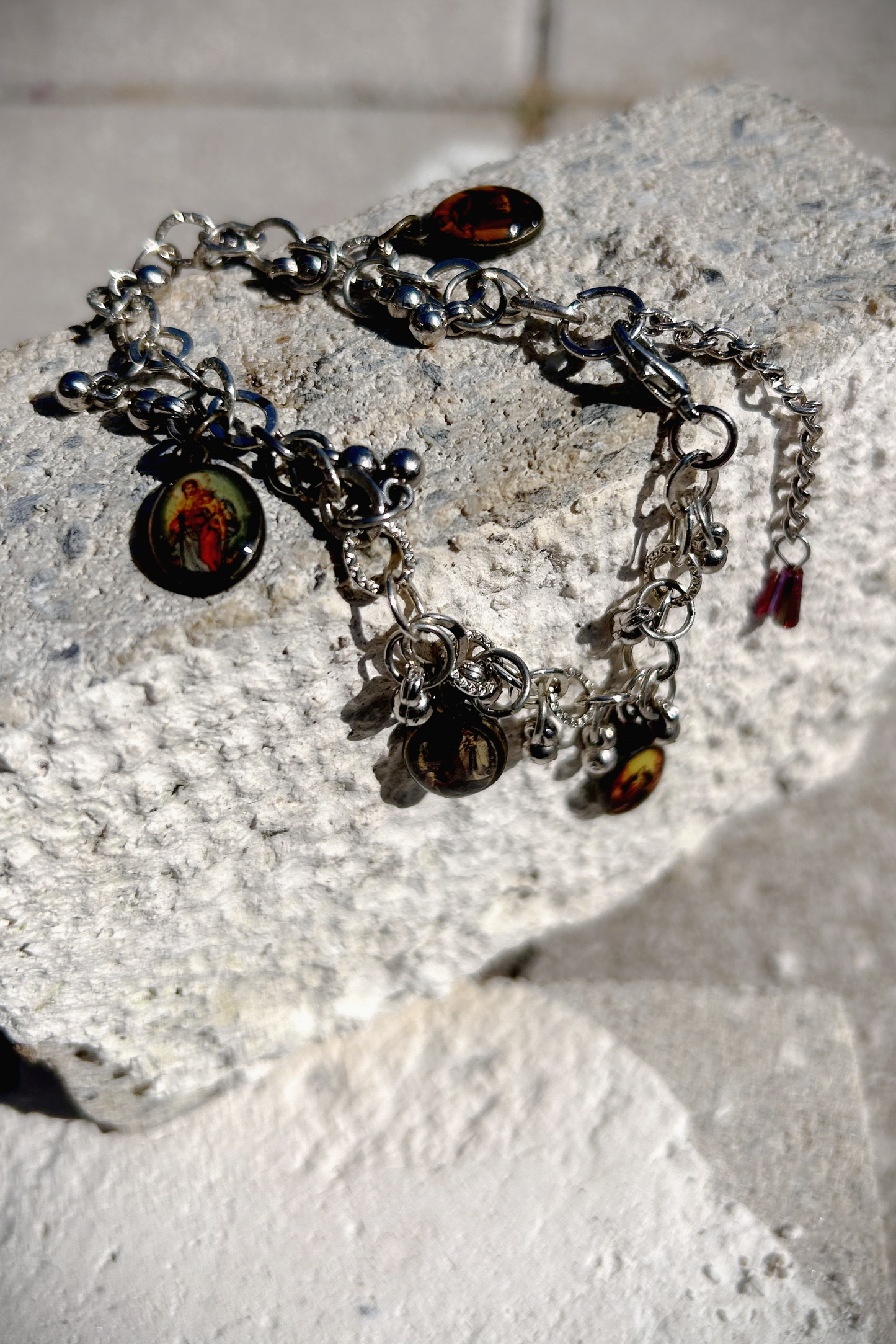 Bracelet recyclé d'un collier, agrémenté de pampilles indiennes, et de médailles religieuses de Saints. J'ai accroché une perle rouge au fermoir, au Japon le rouge protège contre les mauvais esprits et les maladies. Vu du bracelet posé à cheval sur une brique.