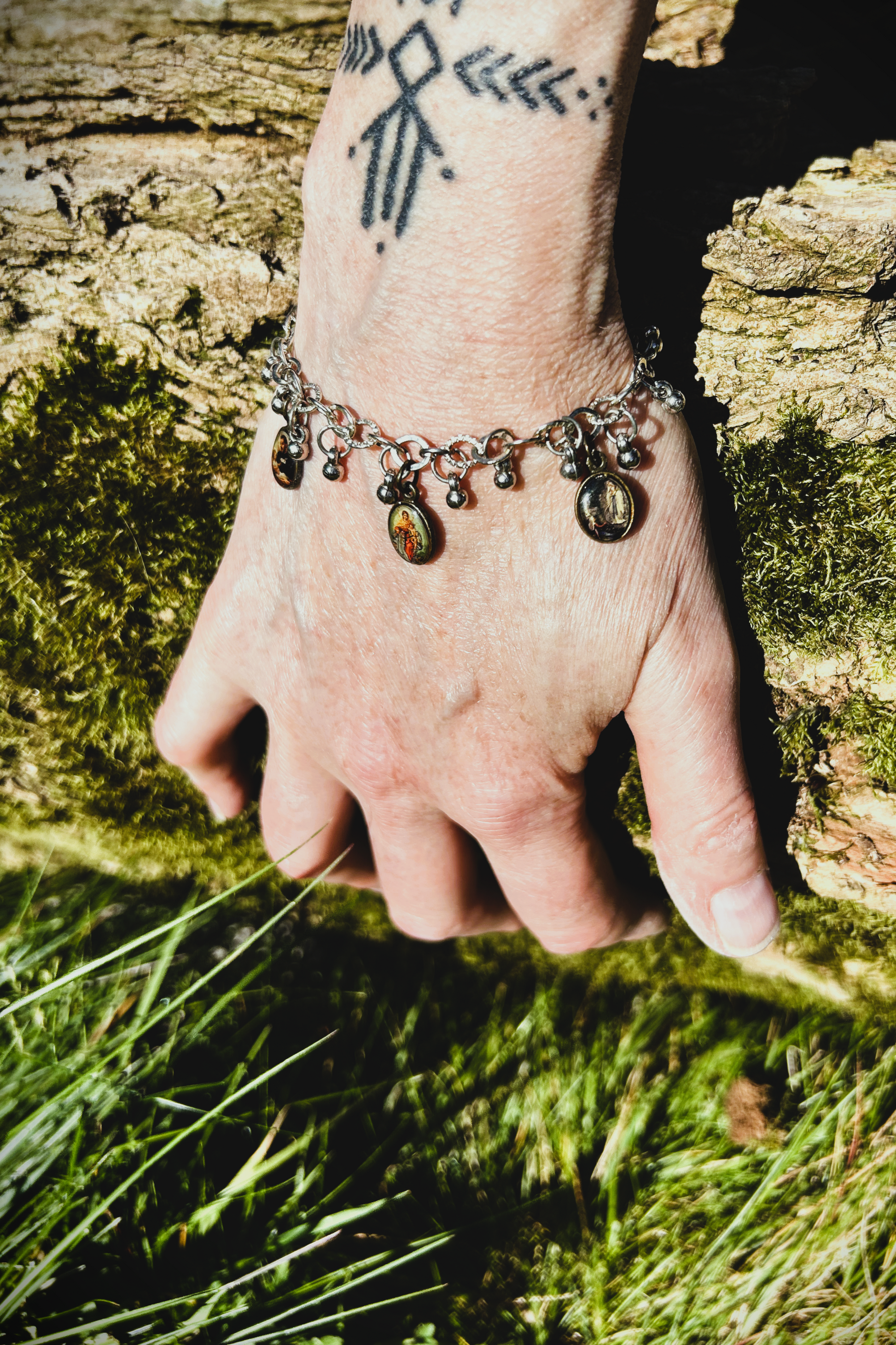 Bracelet recyclé d'un collier, agrémenté de pampilles indiennes, et de médailles religieuses de Saints. J'ai accroché une perle rouge au fermoir, au Japon le rouge protège contre les mauvais esprits et les maladies. Vue du bracelet porté, la main posé sur un tronc d'arbre au soleil.