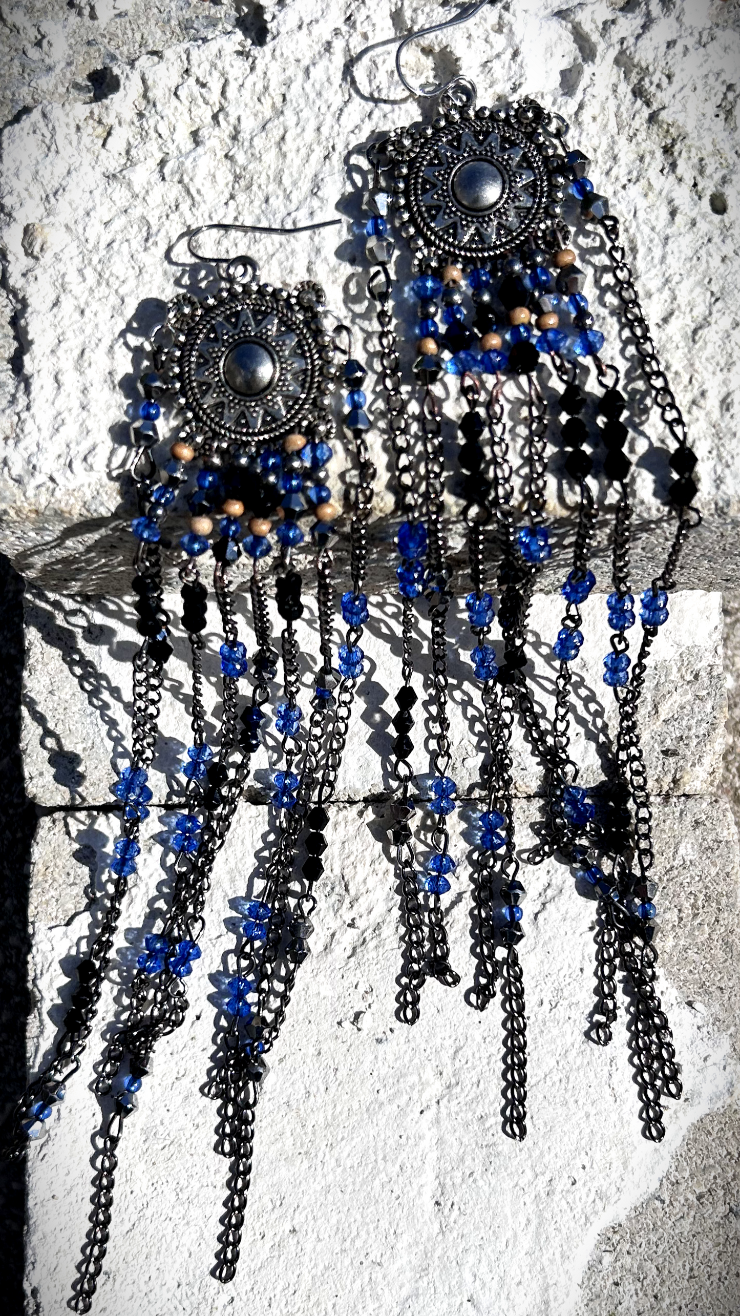 Boucles réalisées comme une évocation des tribus amérindiennes des États-Unis. Pour cela, j’ai récupéré deux maillons de bracelet, que j’ai sciés, limés, émerisés et percés afin d’y suspendre, sur des tiges inox, de longues franges mêlant chaînettes et perles.  Ces franges dans les tons bleutés et noirs en symétrie inversée, apportent un équilibre parfait, et le choix de laisser le premier clou de tige libre permet un mouvement libre à chaque mouvement de tête.