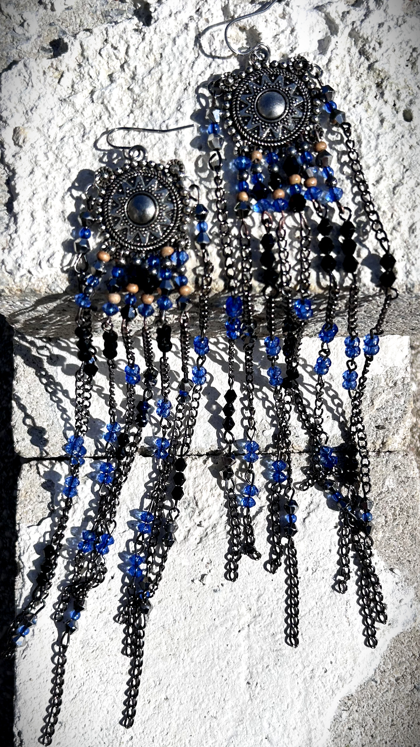 Boucles réalisées comme une évocation des tribus amérindiennes des États-Unis. Pour cela, j’ai récupéré deux maillons de bracelet, que j’ai sciés, limés, émerisés et percés afin d’y suspendre, sur des tiges inox, de longues franges mêlant chaînettes et perles.  Ces franges dans les tons bleutés et noirs en symétrie inversée, apportent un équilibre parfait, et le choix de laisser le premier clou de tige libre permet un mouvement libre à chaque mouvement de tête.