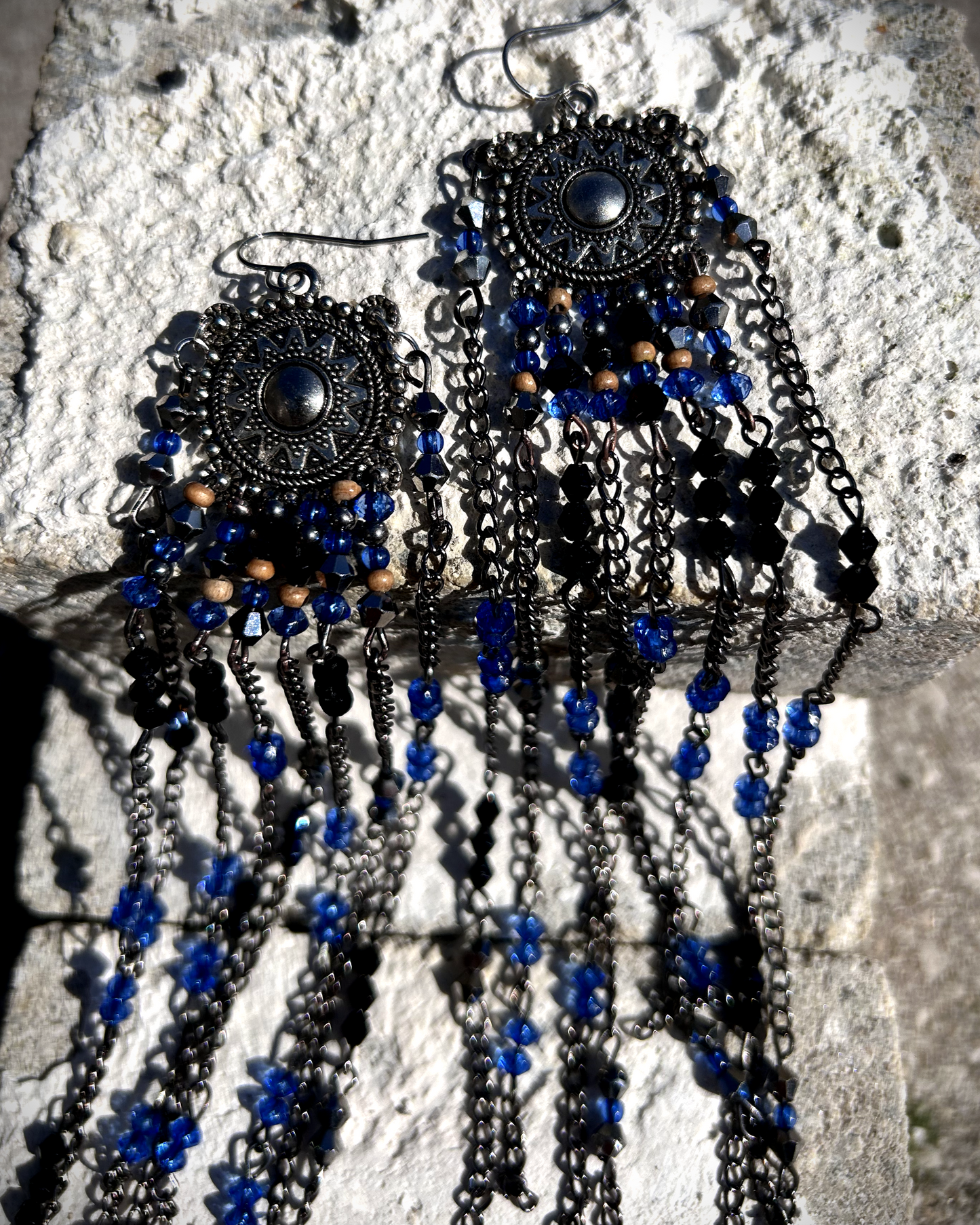 Boucles réalisées comme une évocation des tribus amérindiennes des États-Unis. Pour cela, j’ai récupéré deux maillons de bracelet, que j’ai sciés, limés, émerisés et percés afin d’y suspendre, sur des tiges inox, de longues franges mêlant chaînettes et perles.  Ces franges dans les tons bleutés et noirs en symétrie inversée, apportent un équilibre parfait, et le choix de laisser le premier clou de tige libre permet un mouvement libre à chaque mouvement de tête.