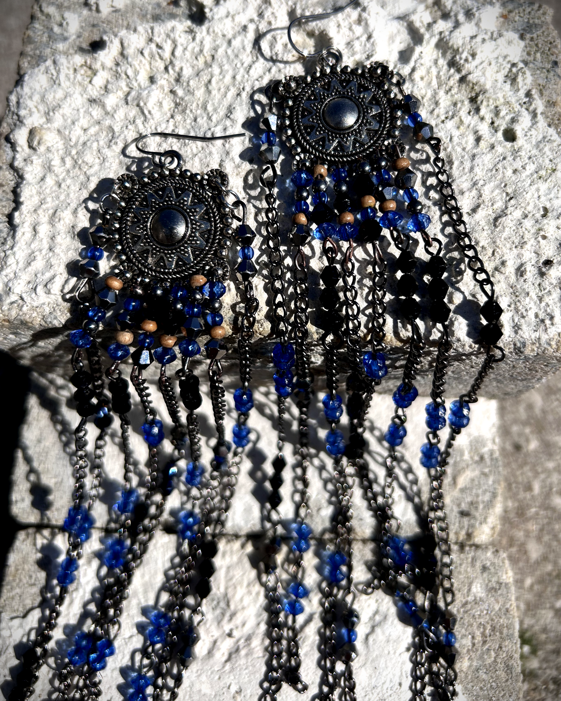 Boucles réalisées comme une évocation des tribus amérindiennes des États-Unis. Pour cela, j’ai récupéré deux maillons de bracelet, que j’ai sciés, limés, émerisés et percés afin d’y suspendre, sur des tiges inox, de longues franges mêlant chaînettes et perles.  Ces franges dans les tons bleutés et noirs en symétrie inversée, apportent un équilibre parfait, et le choix de laisser le premier clou de tige libre permet un mouvement libre à chaque mouvement de tête.