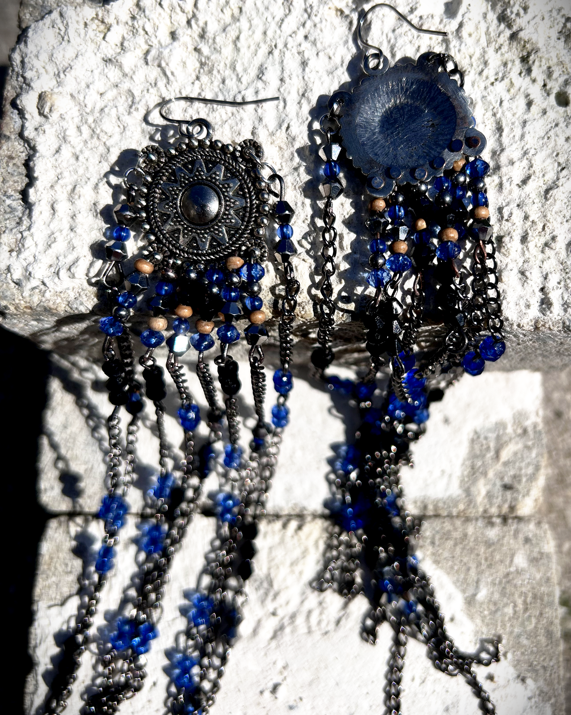 Boucles  évoquant les tribus amérindiennes des États-Unis. Pour cela, j’ai récupéré deux maillons de bracelet, que j’ai sciés, limés, émerisés et percés afin d’y suspendre, sur des tiges inox, de longues franges mêlant chaînettes et perles.  Ces franges dans les tons bleutés et noirs en symétrie inversée, apportent un équilibre parfait. Vue faces avant et arrière.