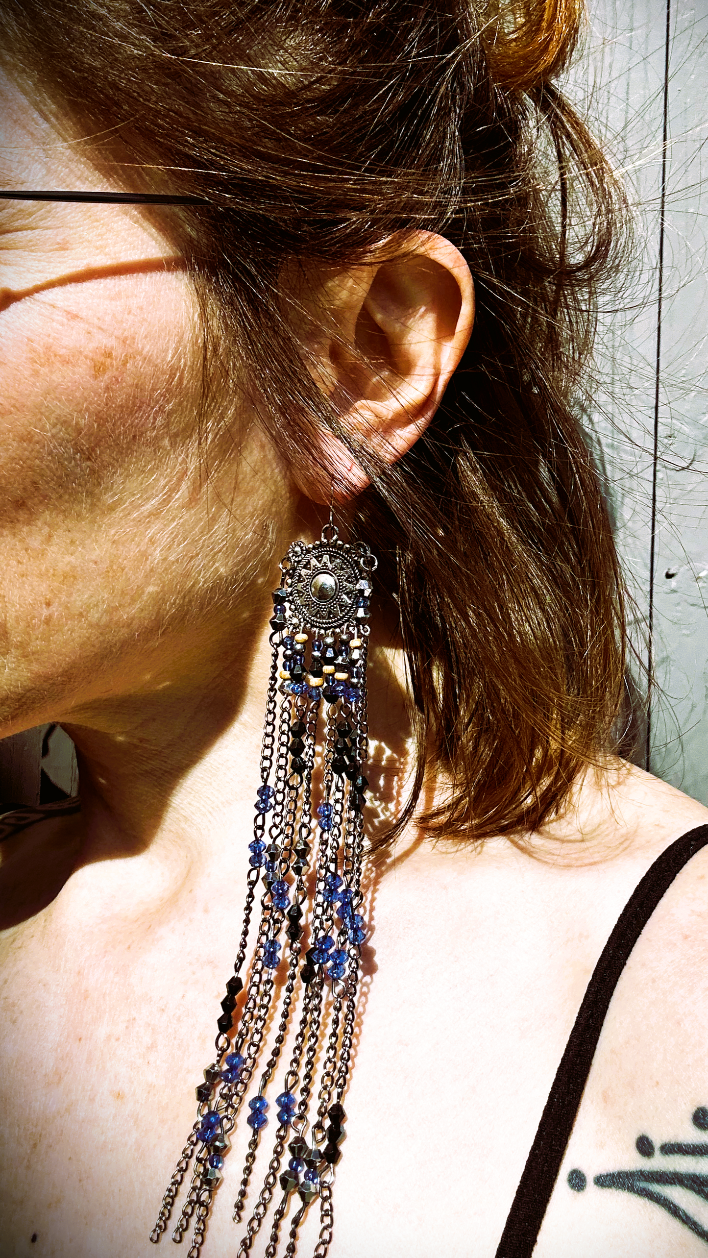 Boucles  évoquant les tribus amérindiennes des États-Unis. Pour cela, j’ai récupéré deux maillons de bracelet, que j’ai sciés, limés, émerisés et percés afin d’y suspendre, sur des tiges inox, de longues franges mêlant chaînettes et perles.  Ces franges dans les tons bleutés et noirs en symétrie inversée, apportent un équilibre parfait. Vue des boucles portées au soleil