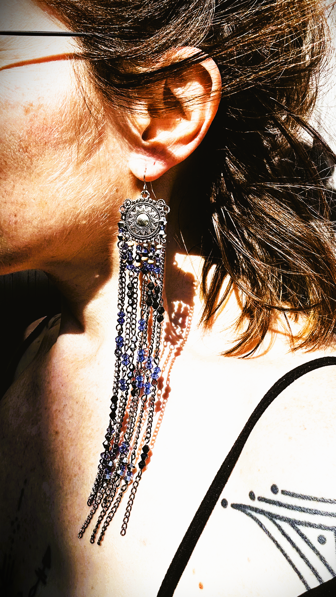 Boucles  évoquant les tribus amérindiennes des États-Unis. Pour cela, j’ai récupéré deux maillons de bracelet, que j’ai sciés, limés, émerisés et percés afin d’y suspendre, sur des tiges inox, de longues franges mêlant chaînettes et perles.  Ces franges dans les tons bleutés et noirs en symétrie inversée, apportent un équilibre parfait. Vue des boucles portées au soleil
