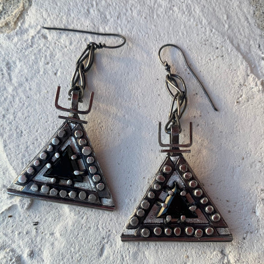 Boucles d'oreilles en argent, baptisées 'Béliers Noirs', inspirées par le symbole berbère du bélier. Elles mesurent 7,5 centimètres de hauteur et sont dotées d'un chaînage sur lequel est suspendu un triangle orné de billes d'argent écrasées sur ses trois côtés. Au centre de ces triangles, un onyx noir facetté de 1cm de côté est serti. Vue de face.