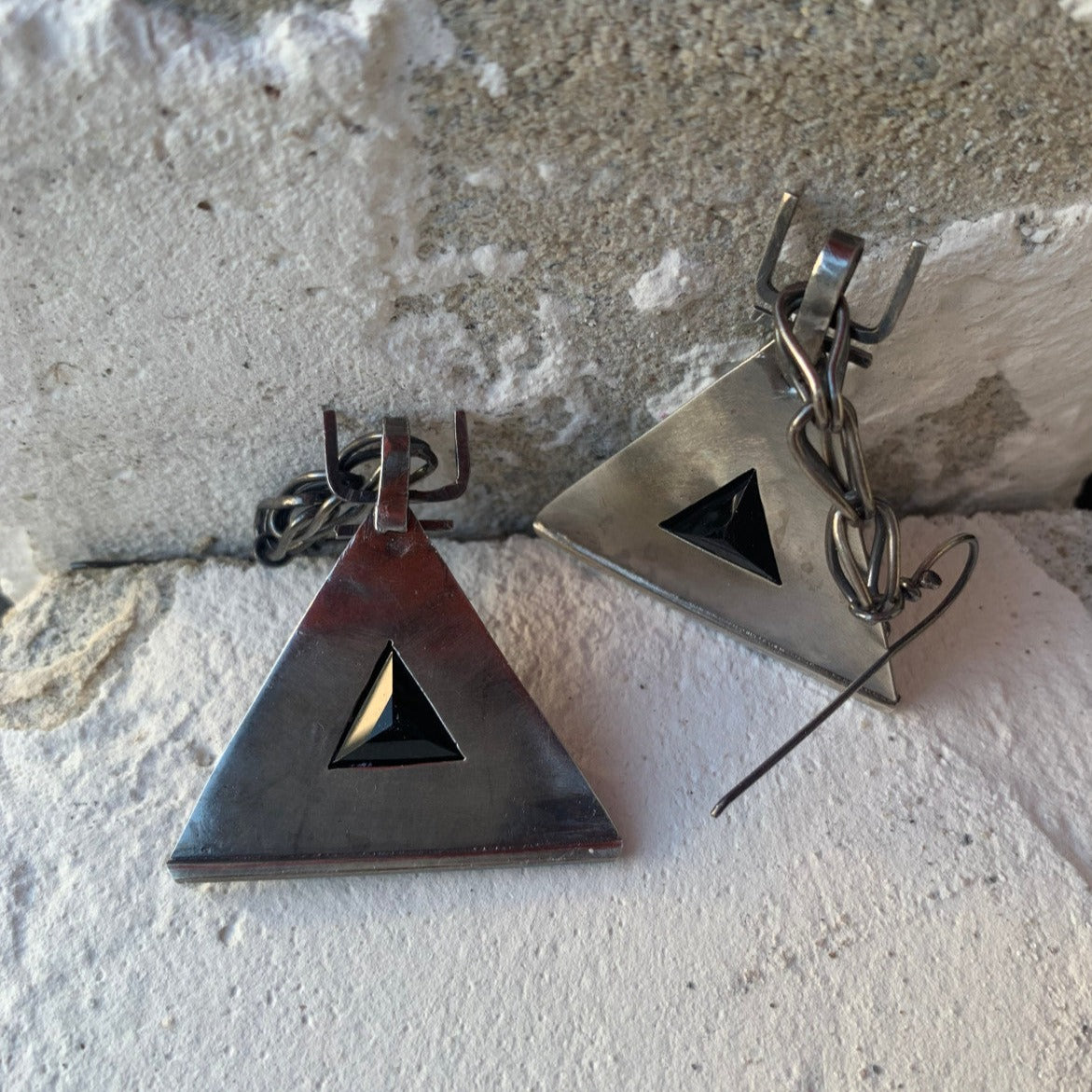 Boucles d'oreilles en argent, baptisées 'Béliers Noirs', inspirées du symbole berbère du bélier. Elles mesurent 7,5 centimètres de hauteur et sont dotées d'un chaînage sur lequel est suspendu un triangle orné de billes d'argent écrasées sur ses trois côtés. Au centre de ces triangles, un onyx noir facetté de 1cm de côté est serti. Vue de dos ici, lisse, laissant apparaitre l'assise de la pierre facettée en pyramide.