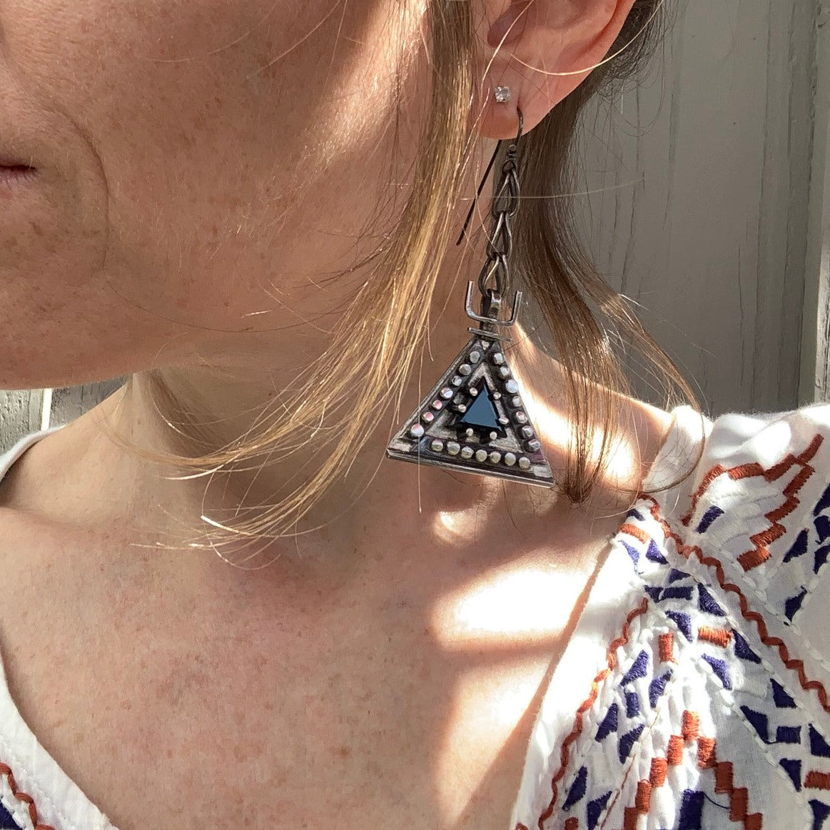 Boucles d'oreilles en argent, baptisées 'Béliers Noirs', inspirées par le symbole berbère du bélier. Elles mesurent 7,5 centimètres de hauteur et sont dotées d'un chaînage sur lequel est suspendu un triangle orné de billes d'argent écrasées sur ses trois côtés. Au centre de ces triangles, un onyx noir facetté de 1cm de côté est serti. Vue de face.