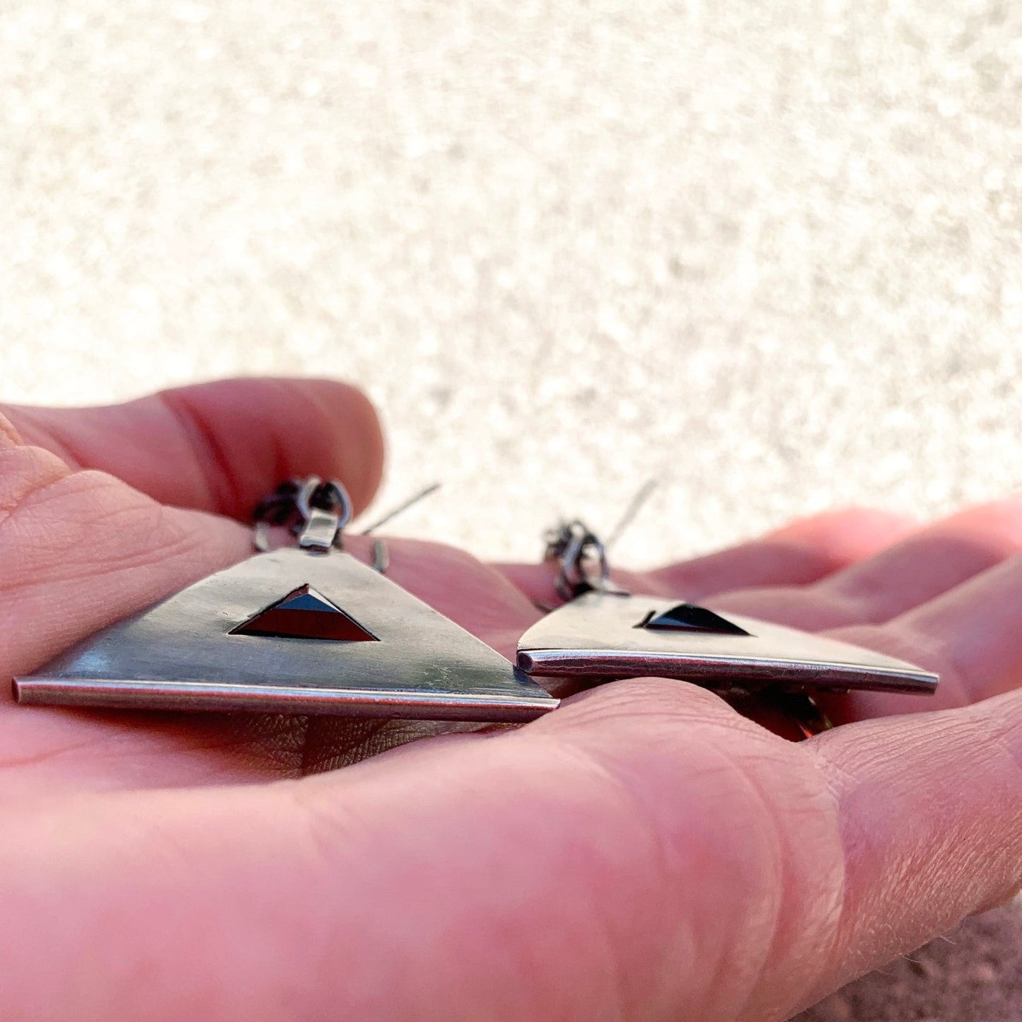 Boucles d'oreilles en argent, baptisées 'Béliers Noirs', inspirées du symbole berbère du bélier. Elles mesurent 7,5 centimètres de hauteur et sont dotées d'un chaînage sur lequel est suspendu un triangle orné de billes d'argent écrasées sur ses trois côtés. Au centre de ces triangles, un onyx noir facetté de 1cm de côté est serti. Vue de dos et de profil ici, lisse, laissant apparaitre l'assise de la pierre facettée en pyramide.