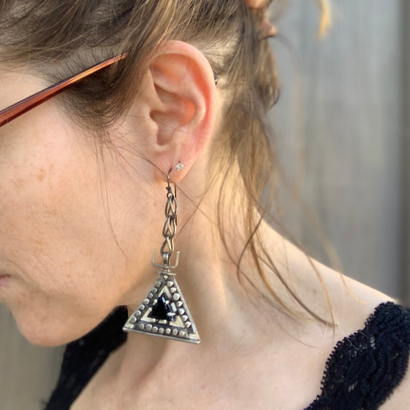 Boucles d'oreilles en argent, baptisées 'Béliers Noirs', inspirées par le symbole berbère du bélier. Elles mesurent 7,5 centimètres de hauteur et sont dotées d'un chaînage sur lequel est suspendu un triangle orné de billes d'argent écrasées sur ses trois côtés. Au centre de ces triangles, un onyx noir facetté de 1cm de côté est serti. Vue de face et portées.