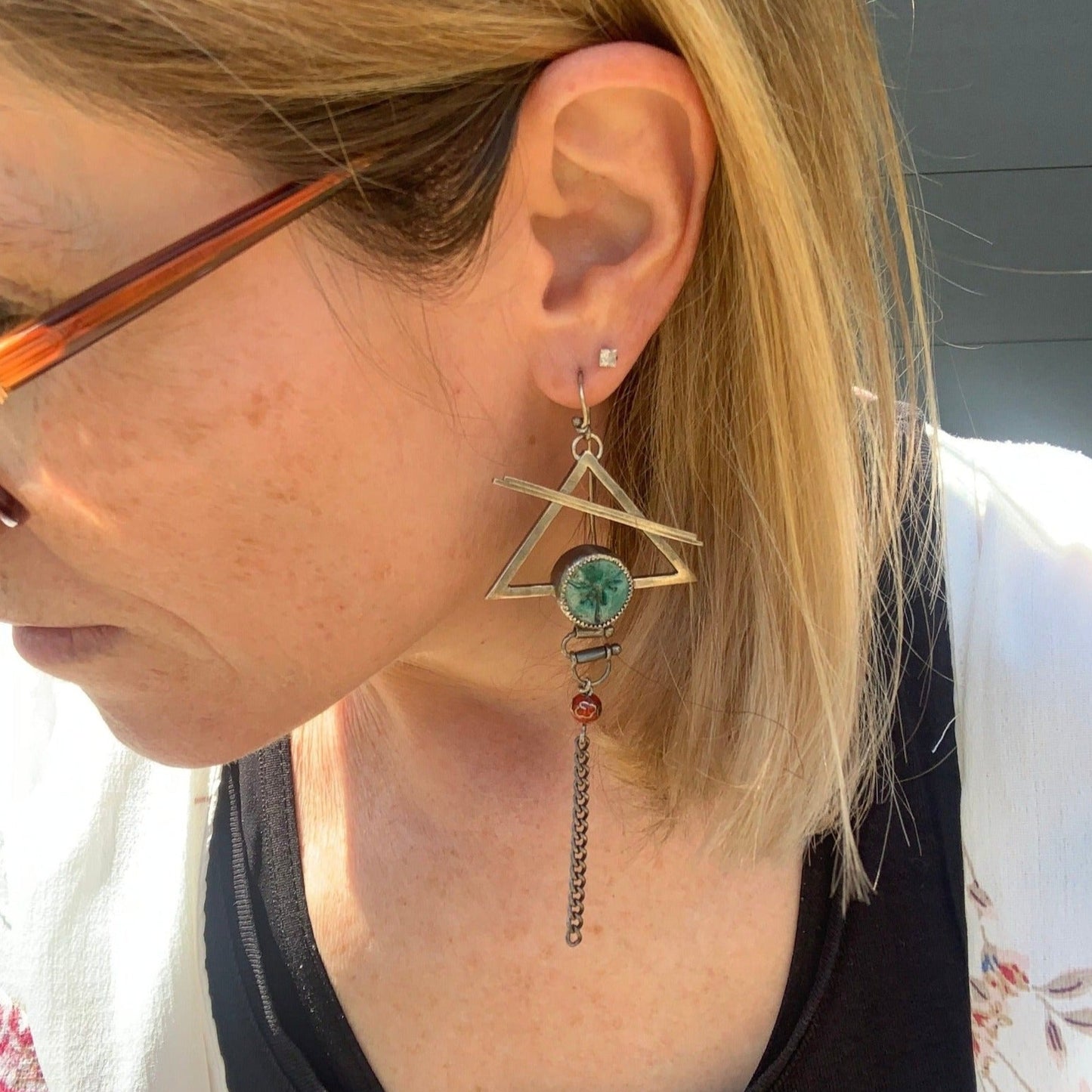 Ces boucles d'oreilles psychédéliques présentent un crochet en argent, au bout duquel est suspendu un triangle évidé traversé par deux barres soudées. Au bas du triangle, un serti en céramique raku irisée ajoute une touche de couleur. Un système de tubes et d'étriers permet de faire bouger la chaîne suspendue, offrant ainsi un effet dynamique. Vue portée.