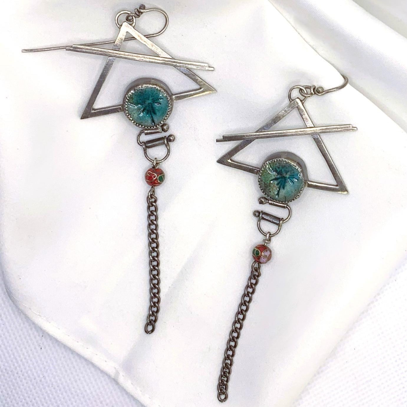 Ces boucles d'oreilles psychédéliques présentent un crochet en argent, au bout duquel est suspendu un triangle évidé traversé par deux barres soudées. Au bas du triangle, un serti en céramique raku irisée ajoute une touche de couleur. Un système de tubes et d'étriers permet de faire bouger la chaîne suspendue, offrant ainsi un effet dynamique. Vue de face. 