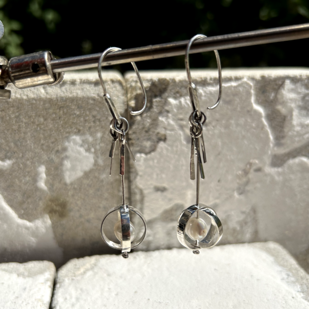 Petites boucles d'oreilles en argent, composé d'un crochet d'oreilles auquel est soudé un maillon d'argent rectangulaire avec coins arrondi. Sur celui-ci se balance une tige qui traverse à sa base deux cercles d'agent qui se croisent symbolisant la terre. Au centre de ces sphères, sur la tige, ont été intégrée sur chaque boucles une perle d'eau de forme irrégulière. Sur le haut de la tige ont été accrochées des petites pampilles illustrant les queues de la comètes.