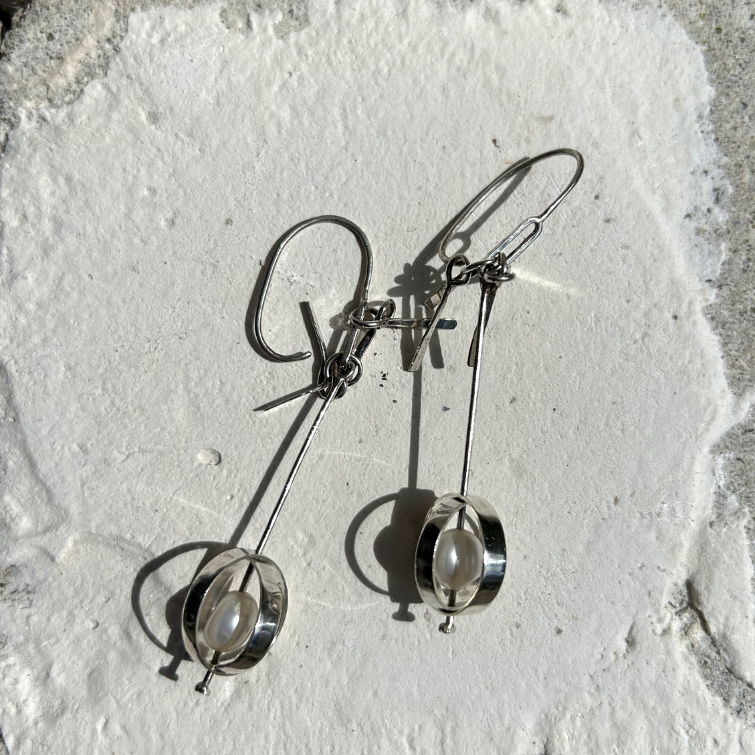 Petites boucles d'oreilles en argent, composé d'un crochet d'oreilles auquel est soudé un maillon d'argent rectangulaire avec coins arrondis. Sur celui-ci se balance une tige qui traverse à sa base deux cercles d'agent qui se croisent symbolisant la terre. Au centre de ces sphères, sur la tige, ont été intégrée sur chaque boucles une perle d'eau de forme irrégulière. Sur le haut de la tige ont été accrochées des petites pampilles illustrant les queues de la comètes. Vue posées.