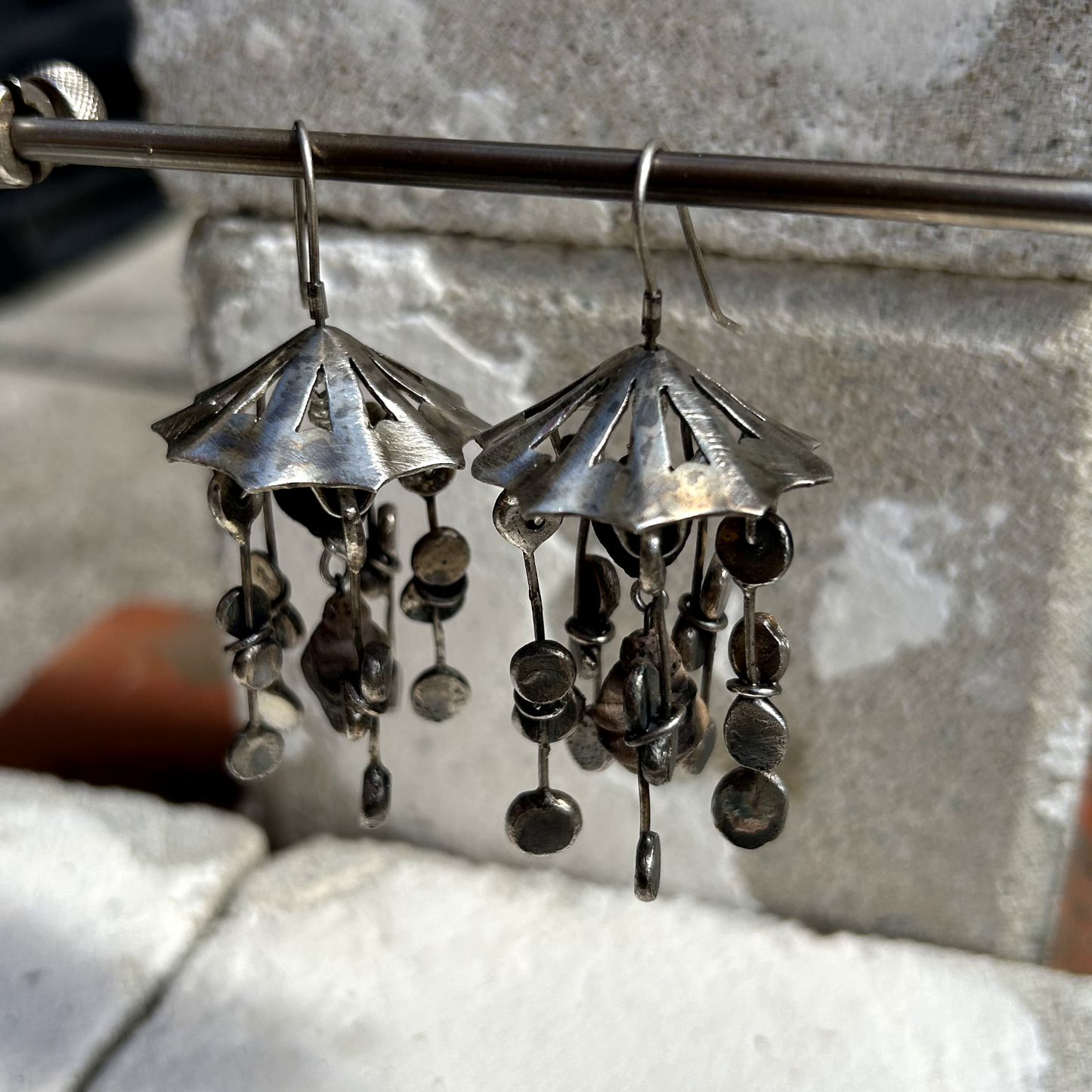 Boucles d'oreilles en argent de 6 cm de hauteur. Crochets d'oreilles en argent. DESIGN : ombrelle en argent sur le haut, ajourée par des petits triangles. À chaque pointe de l'ombrelle est suspendu un assemblage de fils d'argent, avec un aplat à chaque bout. Cinq lierres par boucle. Au centre est accrochée une minuscule représentation de la Vénus de Willendorf en céramique.