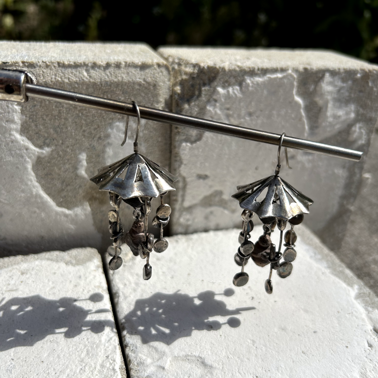 Boucles d'oreilles en argent de 6 cm de hauteur. Crochets d'oreilles en argent. DESIGN : ombrelle en argent sur le haut, ajourée par des petits triangles. À chaque pointe de l'ombrelle est suspendu un assemblage de fils d'argent, avec un aplat à chaque bout. Cinq lierres par boucle. Au centre est accrochée une minuscule représentation de la Vénus de Willendorf en céramique. Vue de coté.