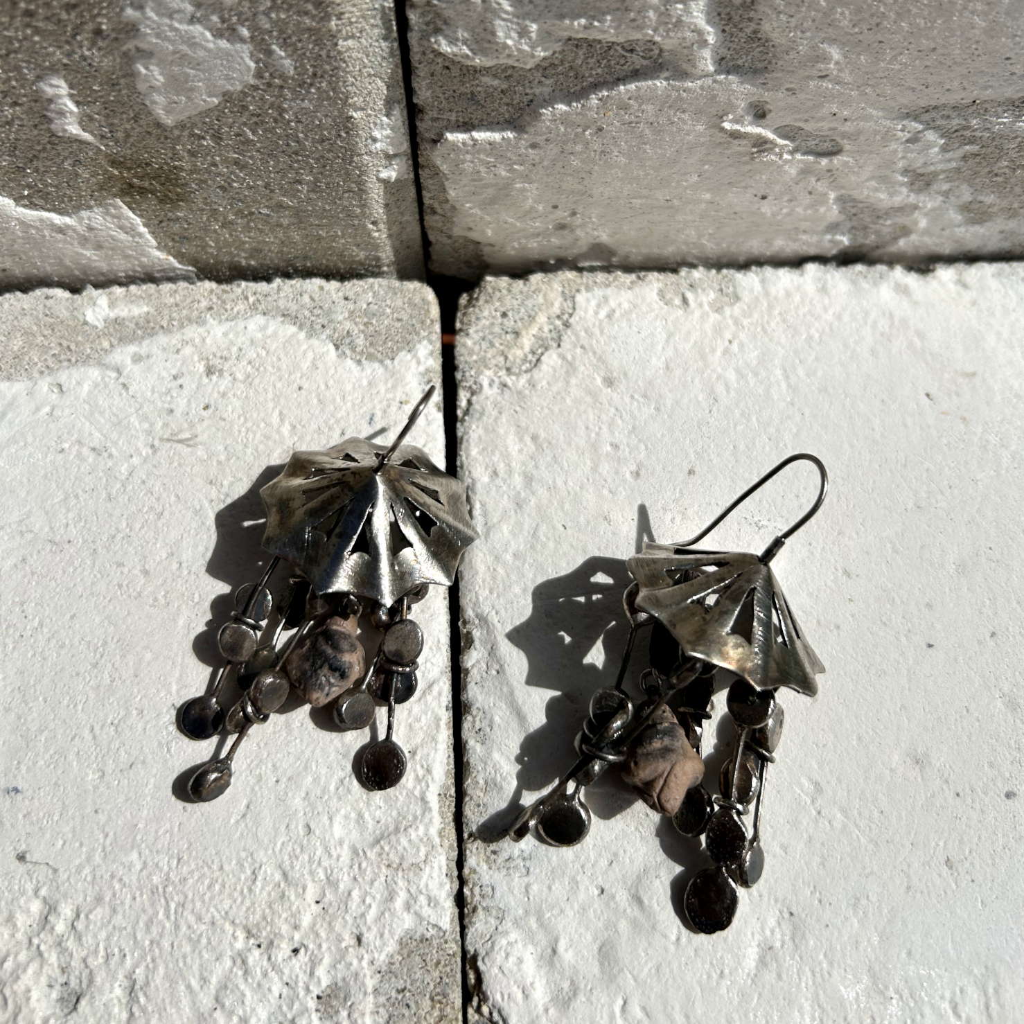 Boucles d'oreilles en argent de 6 cm de hauteur. Crochets d'oreilles en argent. DESIGN : ombrelle en argent sur le haut, ajourée par des petits triangles. À chaque pointe de l'ombrelle est suspendu un assemblage de fils d'argent, avec un aplat à chaque bout. Cinq lierres par boucle. Au centre est accrochée une minuscule représentation de la Vénus de Willendorf en céramique. Vue posées à plat.