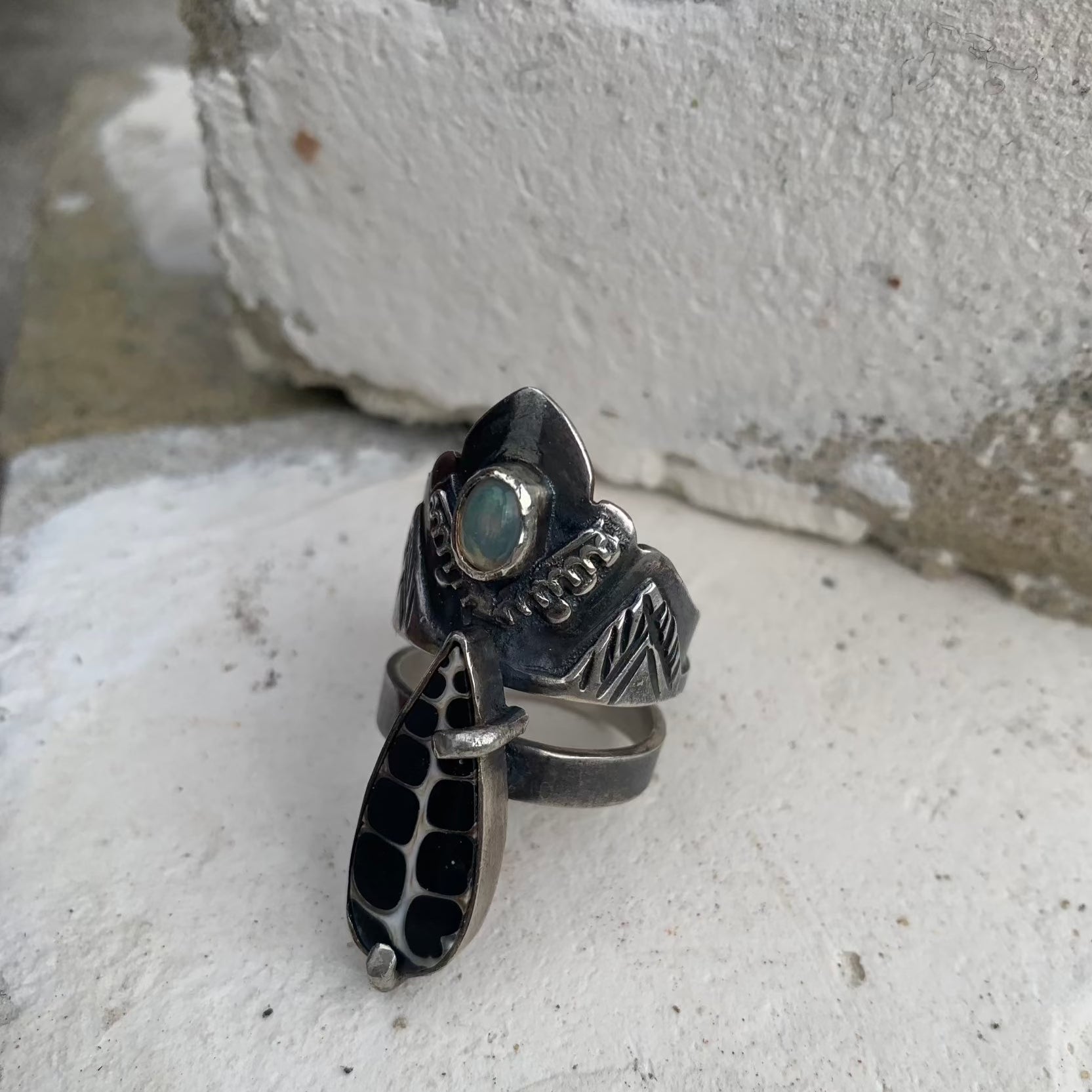 Cette bague en argent se compose de deux joncs distincts soudés. Sur l'un des deux fin est soudé un serti avec un snail fossile sur broches qui relient les deux joncs. Sur le jonc large on retrouve le symbole de l'olivier au centre duquel est serti une petite opale welo représentant une olive. Bague force de résilience et de combativité. Vue de profil.