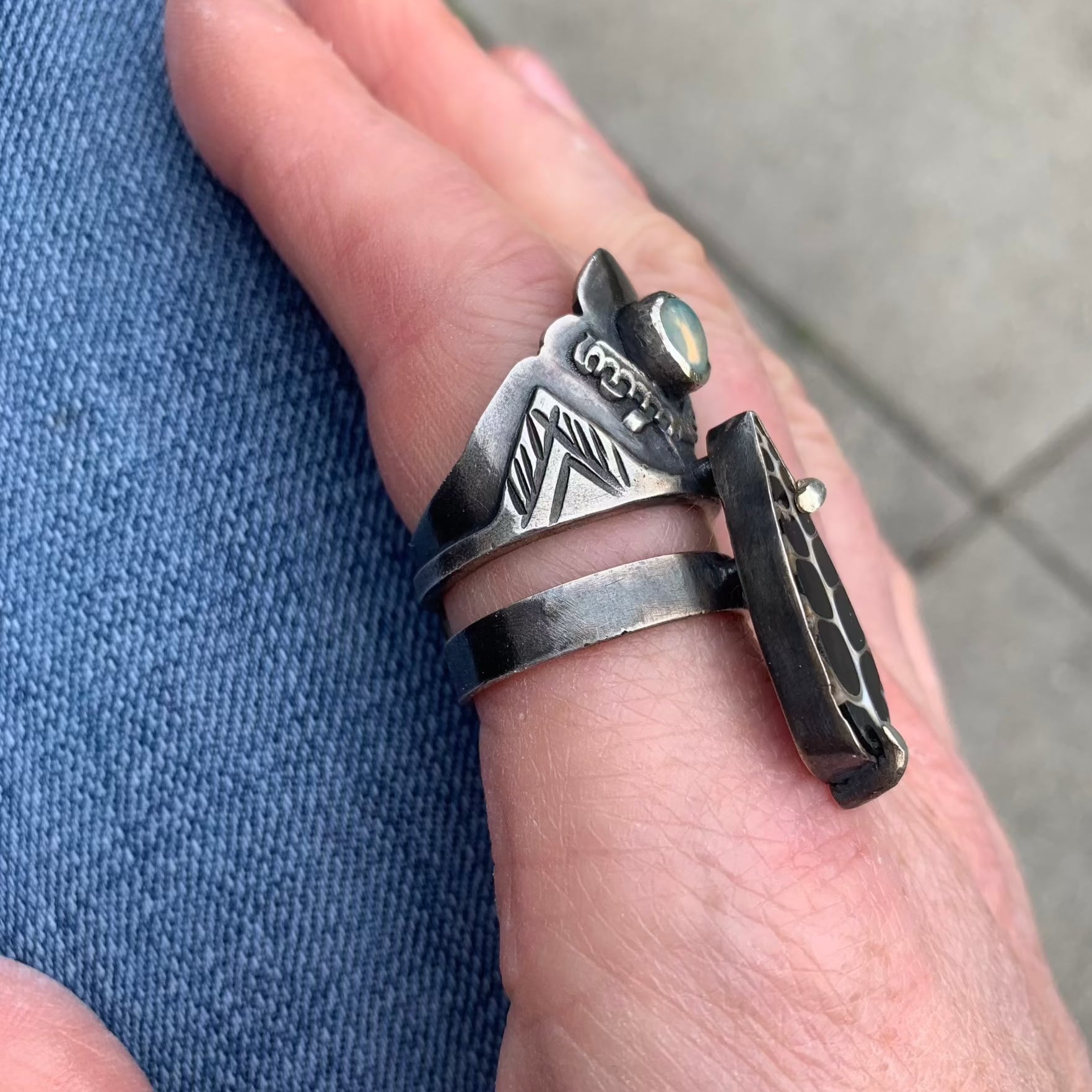 Cette bague en argent se compose de deux joncs distincts. Sur l'un des deux, un serti est soudé, portant un escargot fossile sur des broches qui relient les deux joncs. Sur le jonc large, on retrouve le symbole de l'olivier, au centre duquel est sertie une petite opale Welo représentant une olive. Cette bague incarne la force de résilience et de combativité. Vue de profil.