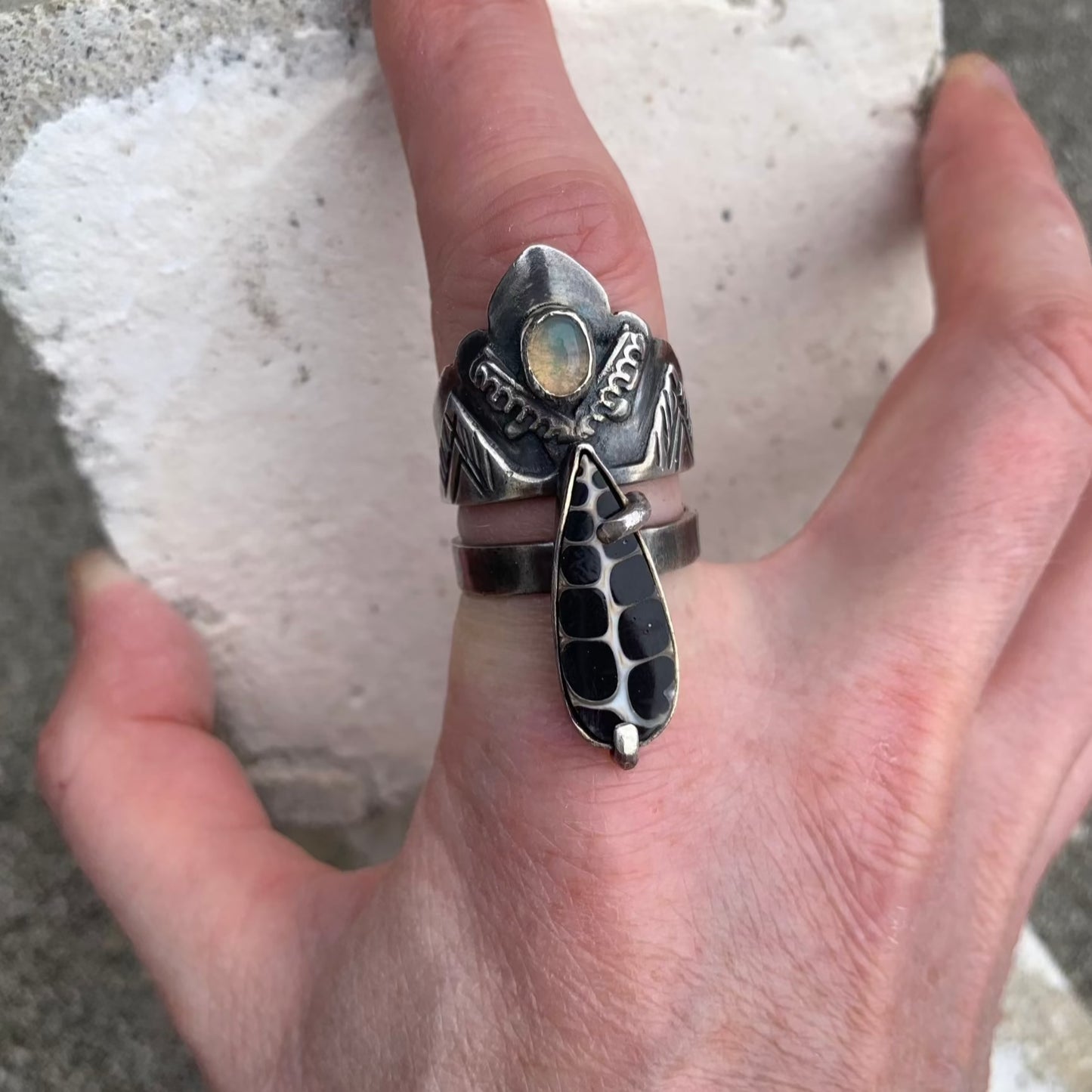 Cette bague en argent se compose de deux joncs distincts. Sur l'un des deux, un serti est soudé, portant un escargot fossile sur des broches qui relient les deux joncs. Sur le jonc large, on retrouve le symbole de l'olivier, au centre duquel est sertie une petite opale Welo représentant une olive. Cette bague incarne la force de résilience et de combativité. Vue de dessus.